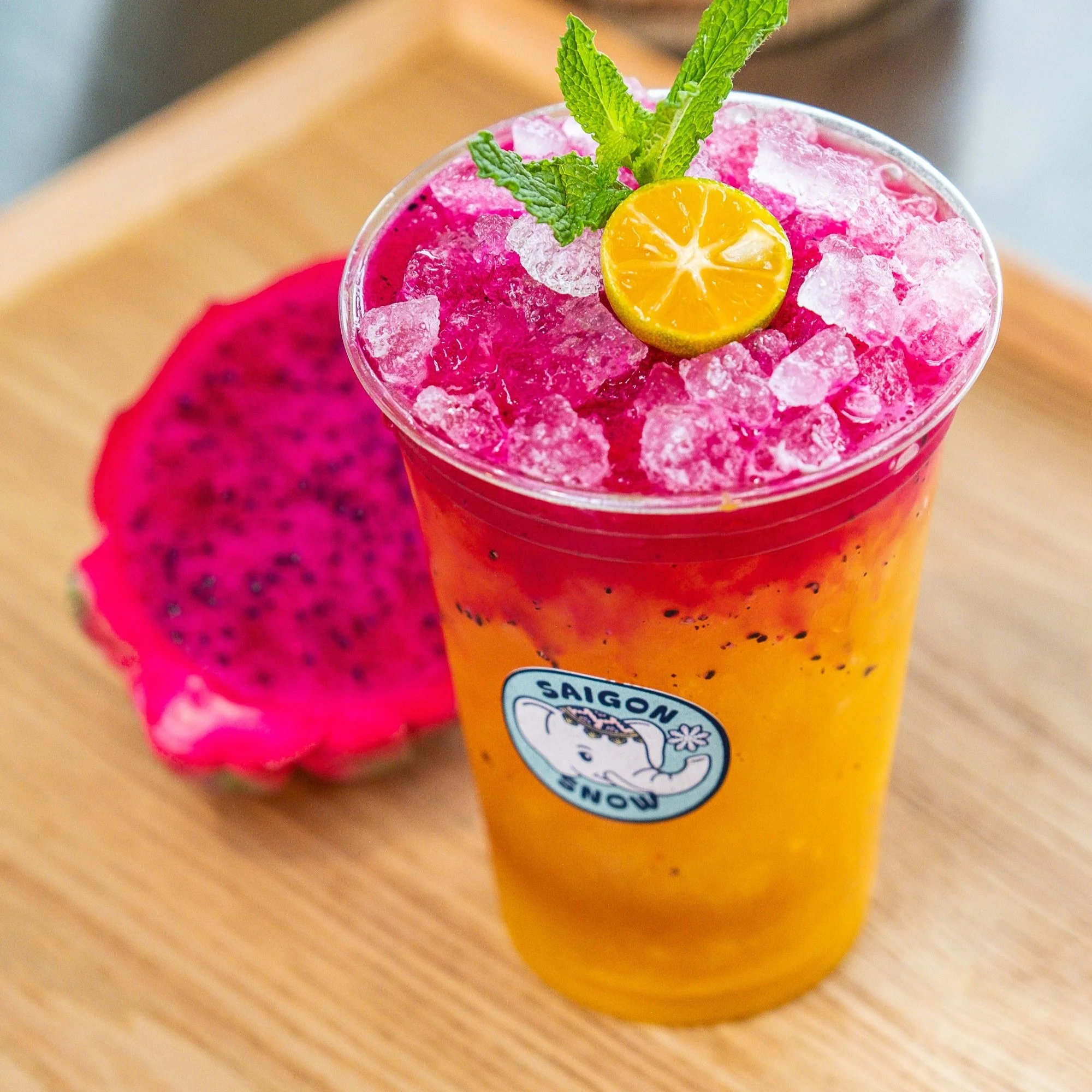 A tall glass of vibrant orange and pink layered fruit drink topped with crushed ice, a small yellow citrus slice, and mint leaves, placed on a wooden surface with a slice of dragon fruit beside it.