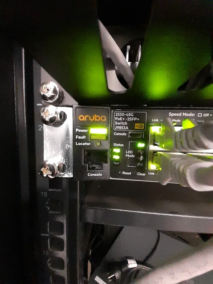 A close-up of a network switch labeled 'aruba' with various indicator lights on and connected to Ethernet cables.