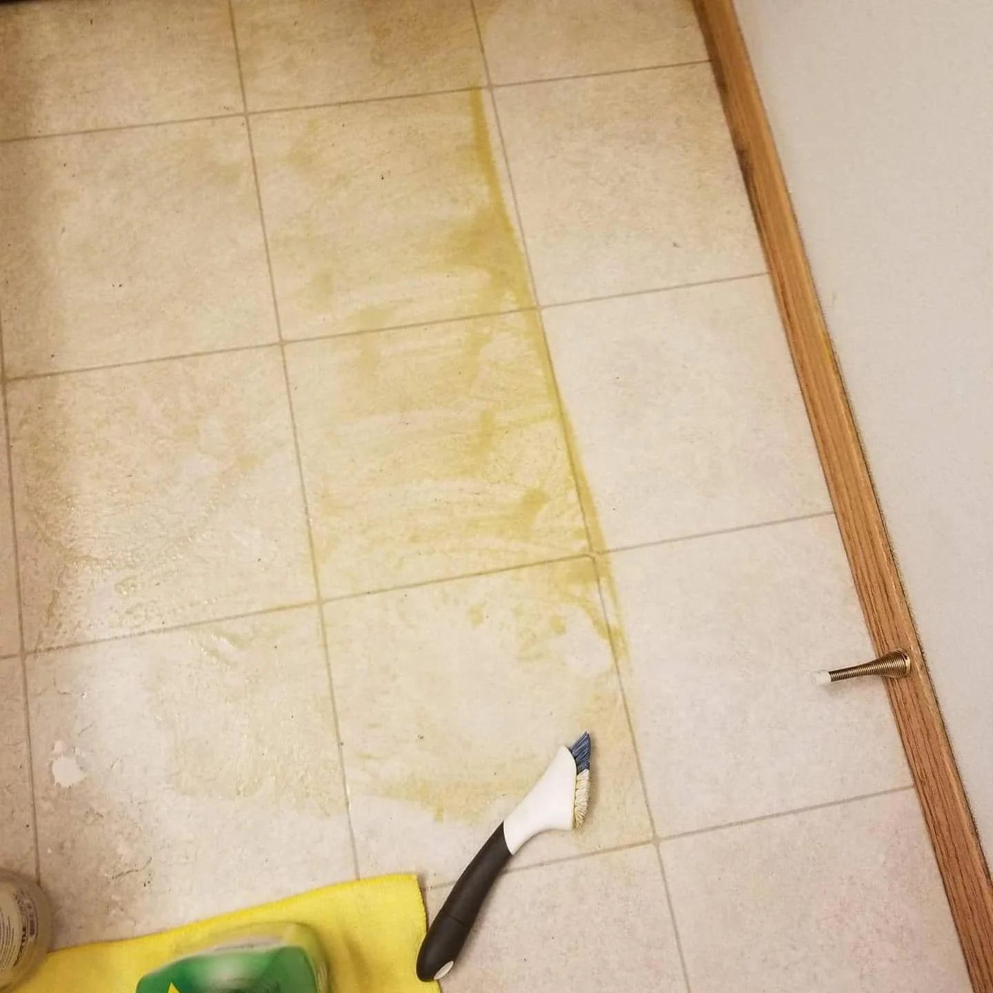 The image shows a tiled floor with major yellowish grime and dirt residue, a grout cleaning tool, a yellow cleaning cloth, and a bottle of cleaning solution, with a wooden baseboard along the edge.