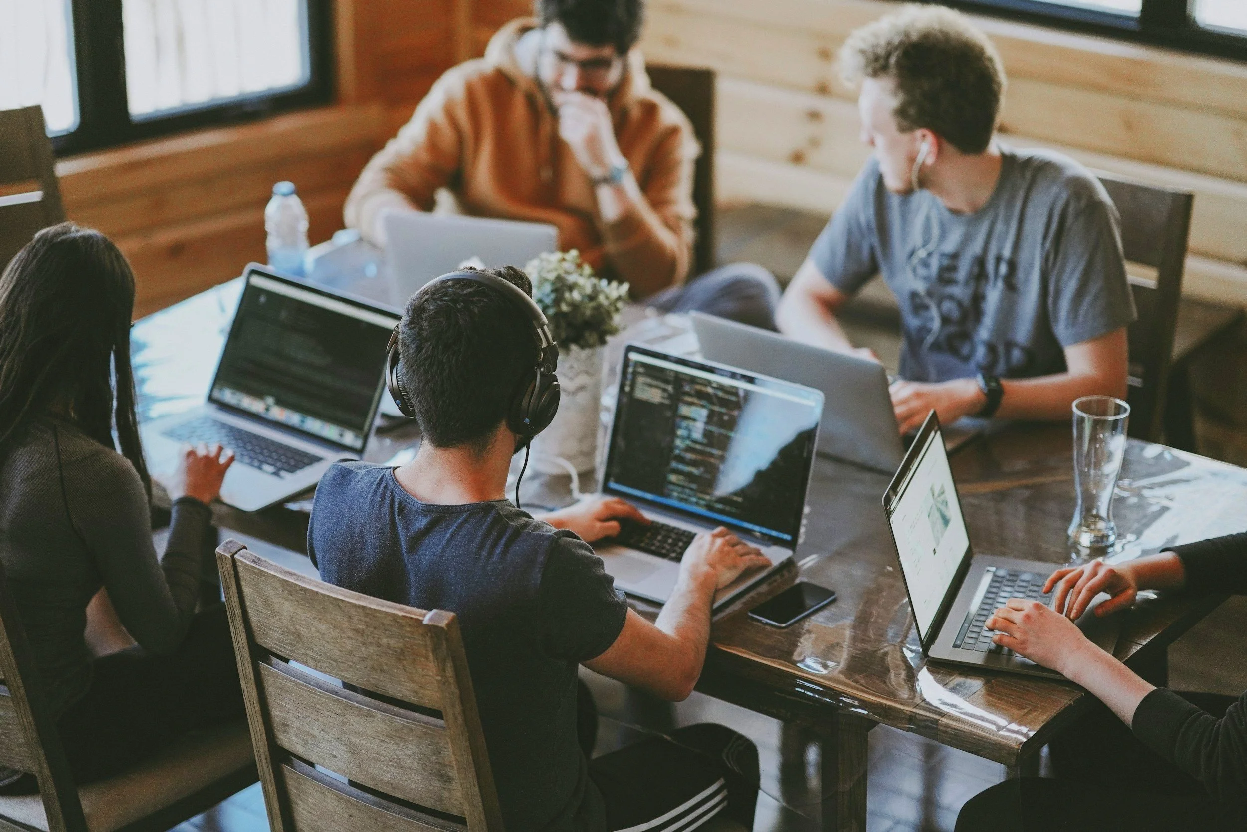 Group of five young adults working together on laptops around a wooden table, some wearing headphones, in a cozy wooden room with large windows.