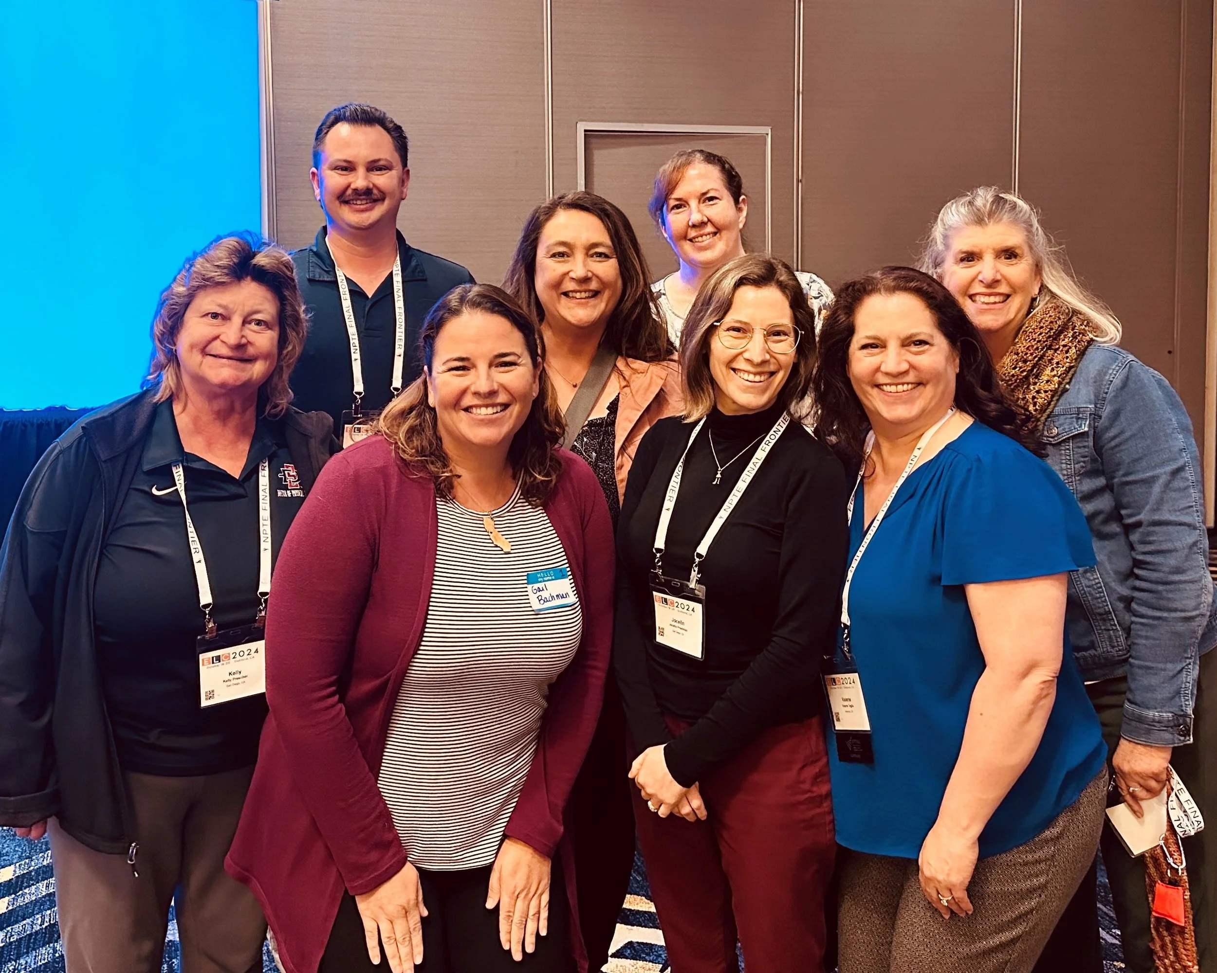 A group of nine smiling women and one man posing together at a conference, wearing name badges and lanyards, standing in front of a conference room backdrop.