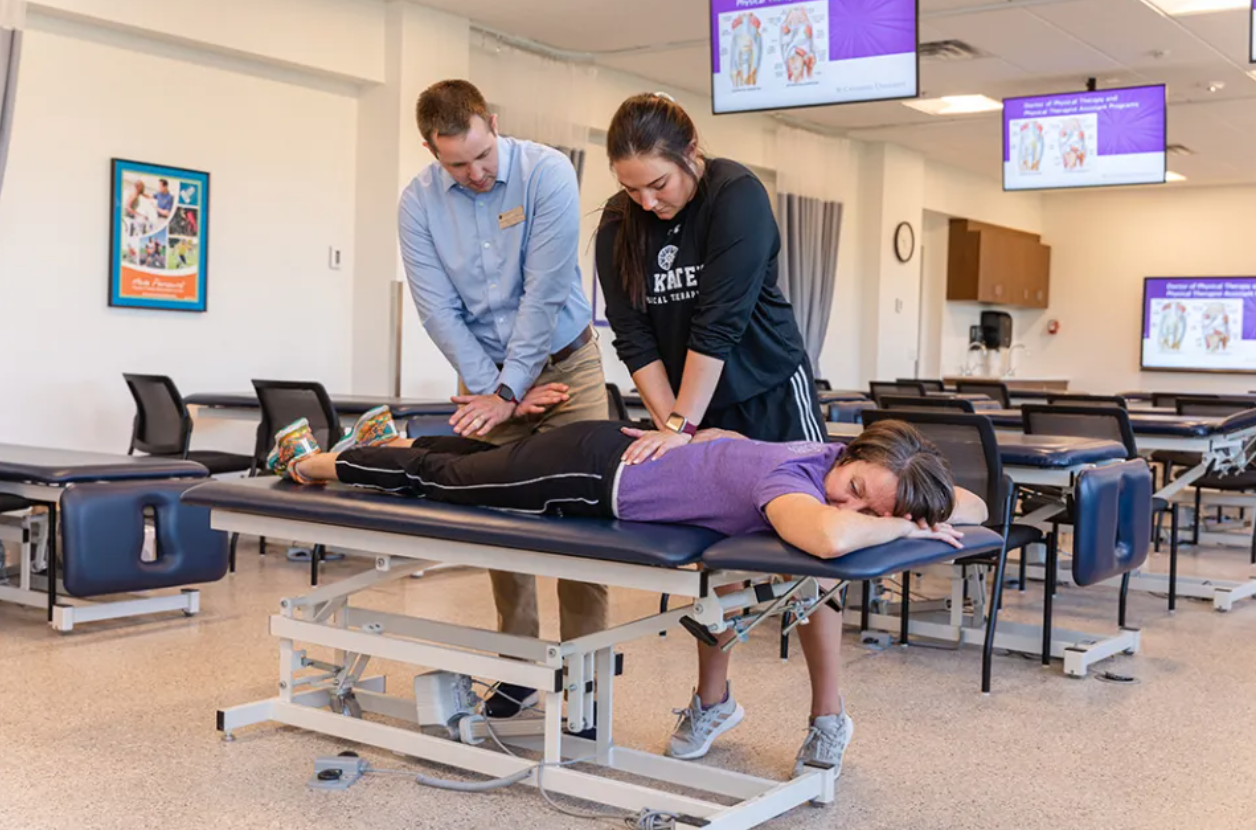 A woman lying on a medical examination table receiving CPR practice from two instructors, a man and a woman, in a classroom setting with multiple television screens displaying educational content in the background.