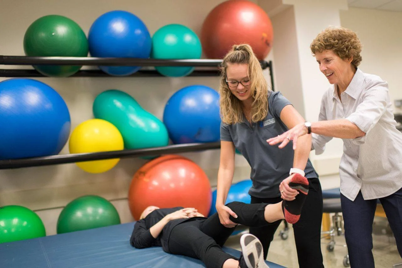 A physical therapist assisting a young woman in stretching or exercising, with a senior woman standing beside them in a fitness or therapy room with colorful exercise balls on shelves in the background.