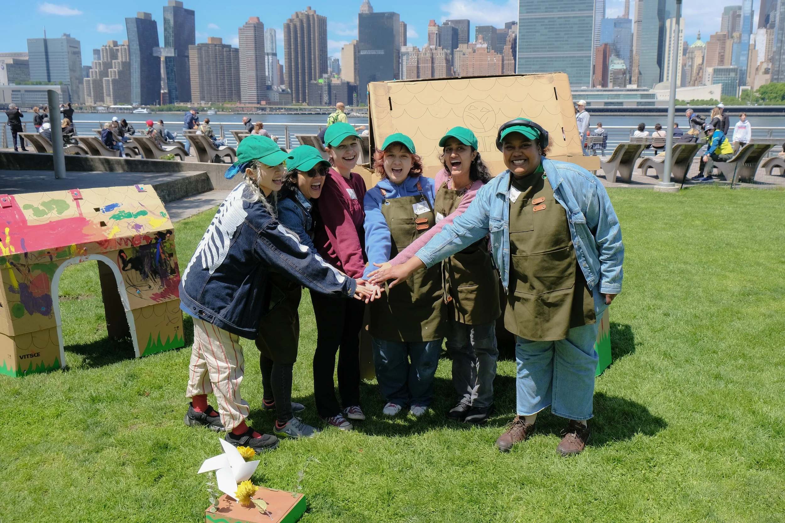 Group of seven diverse women in green caps and casual clothing, smiling and joining hands in a circle outdoors on a grassy area with cardboard structures. In the background, a city skyline with tall buildings and people sitting on benches near a water body.