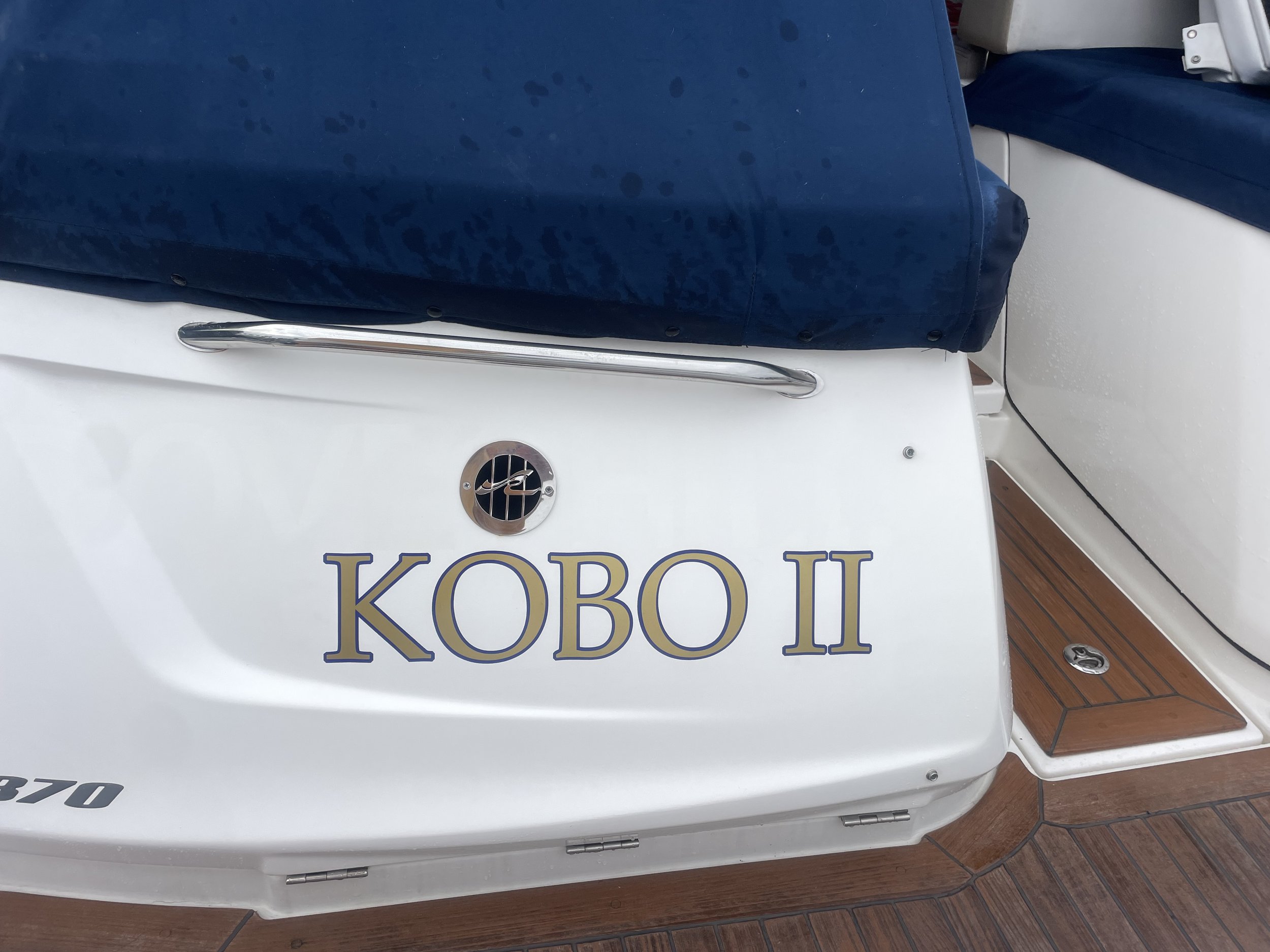 Rear view of a boat with "KOBO II" written on the side, featuring a white hull and navy-blue cover.