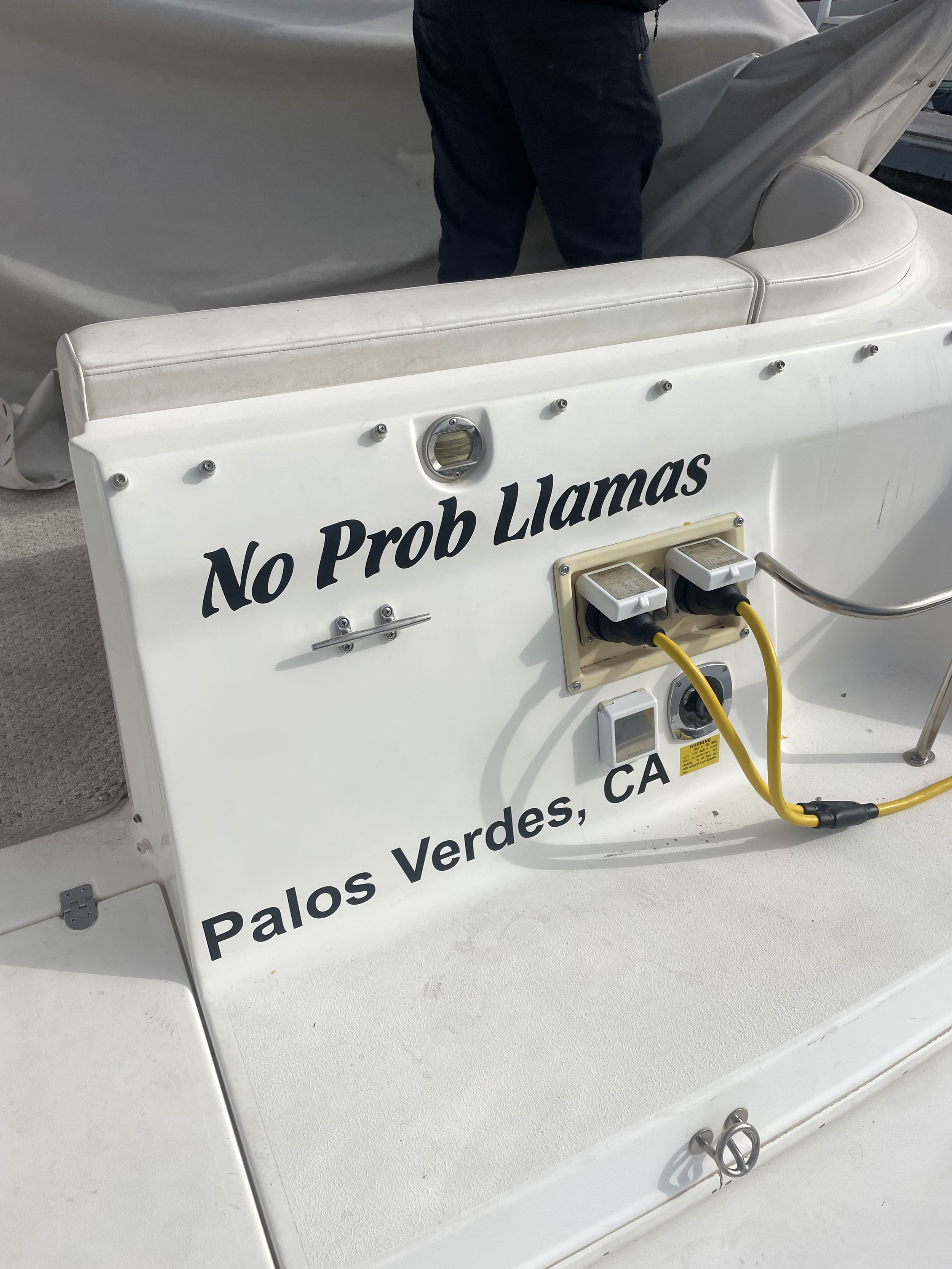Back of a boat with "No Prob Llamas" and "Palos Verdes, CA" written on it, showing a yellow cable plugged into outlets.