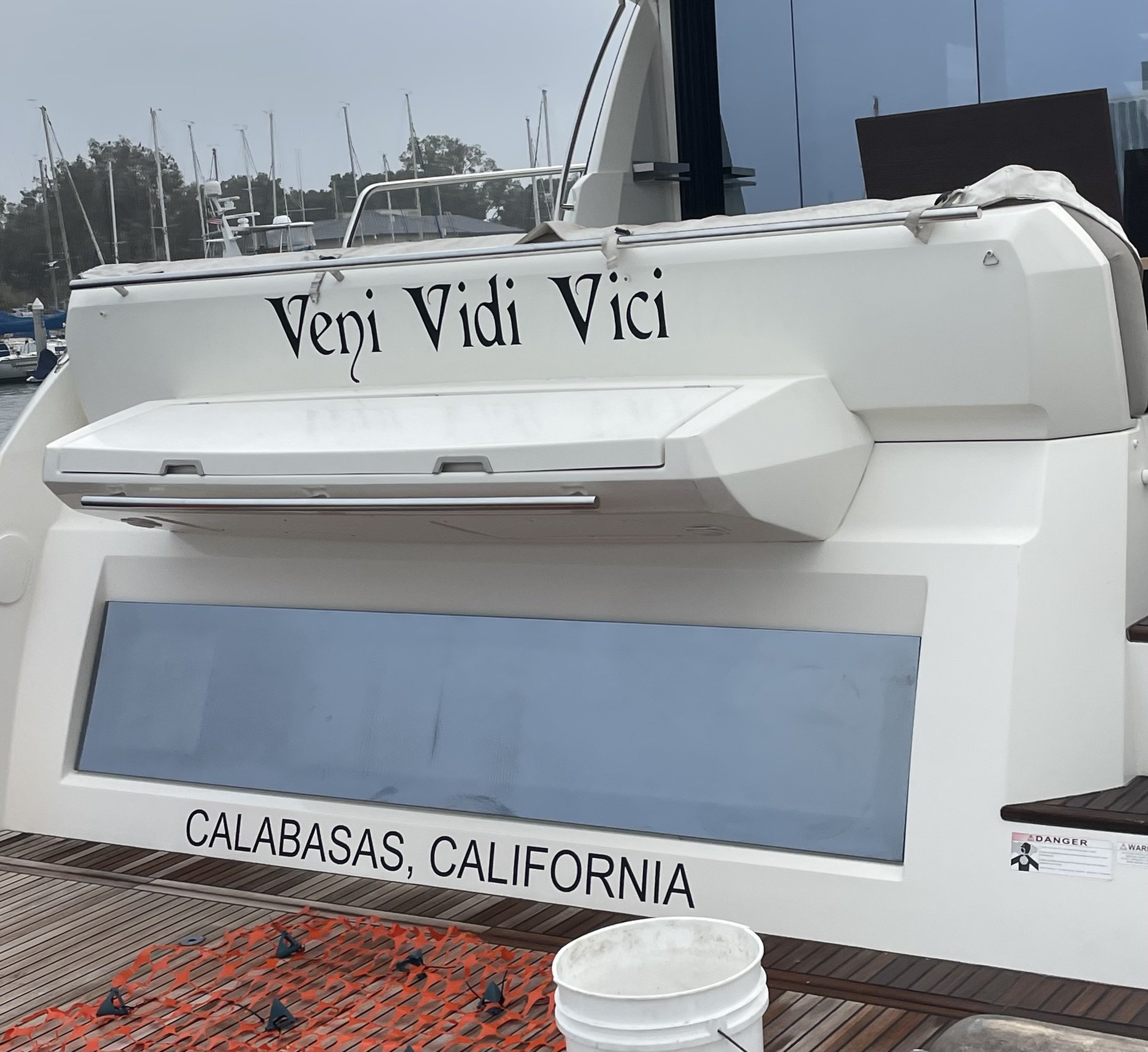 Back of a yacht named 'Veni Vidi Vici' with text stating it is from Calabasas, California, docked with marina background.