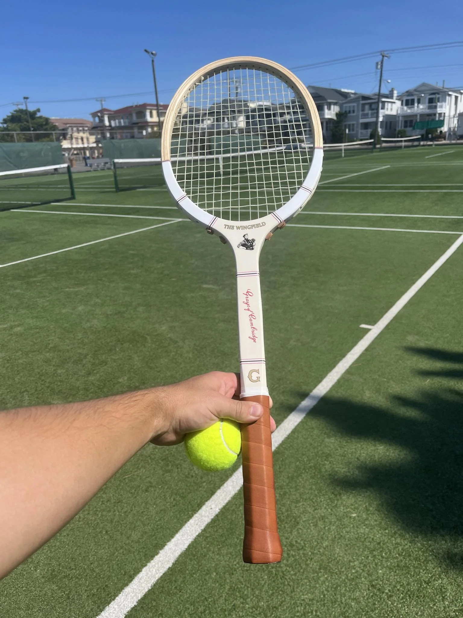 Person holding a Wingfield racket and tennis ball with a row of tennis courts in the background.