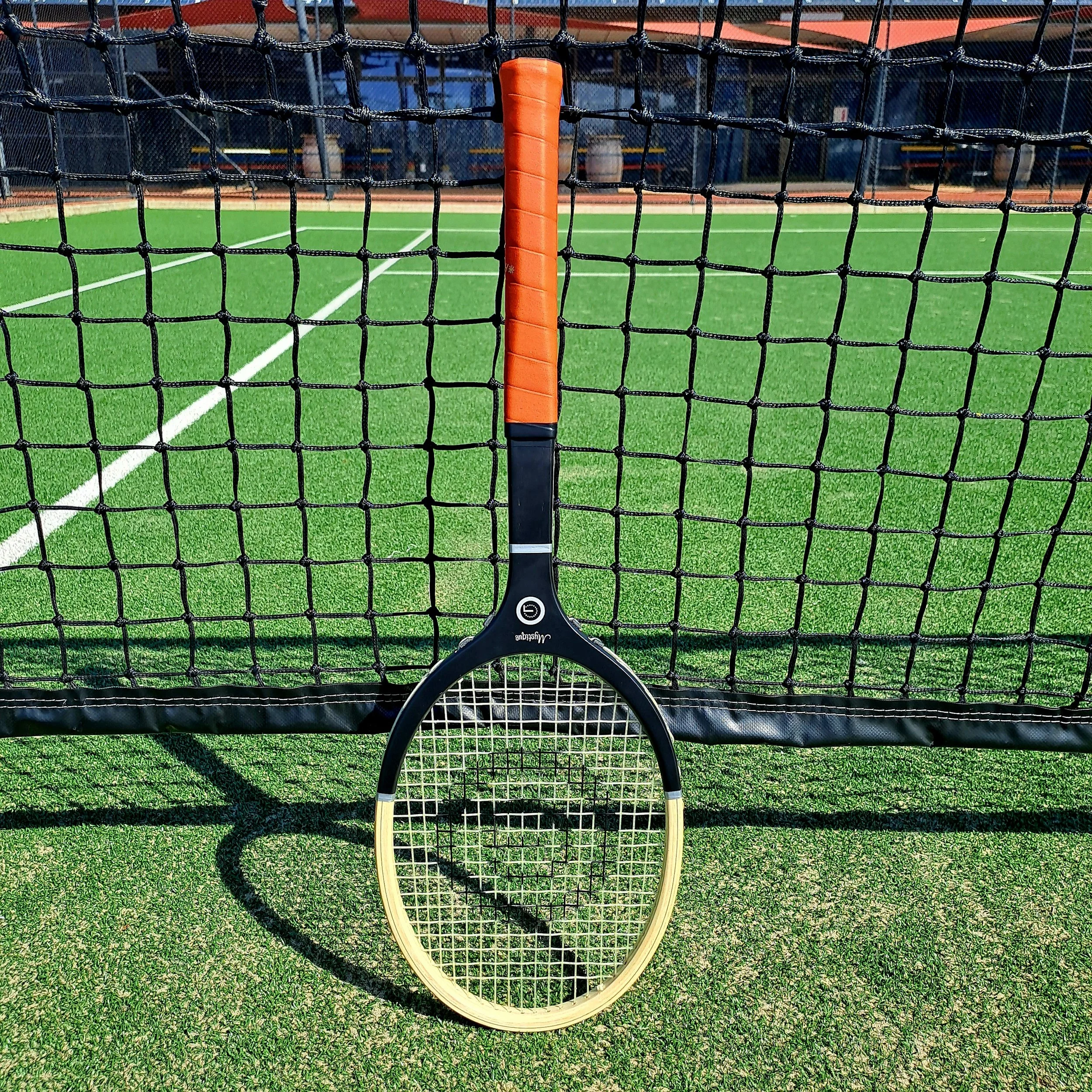 Photo of Grays Mystique resting against net on a grass court with clubhouse in background.