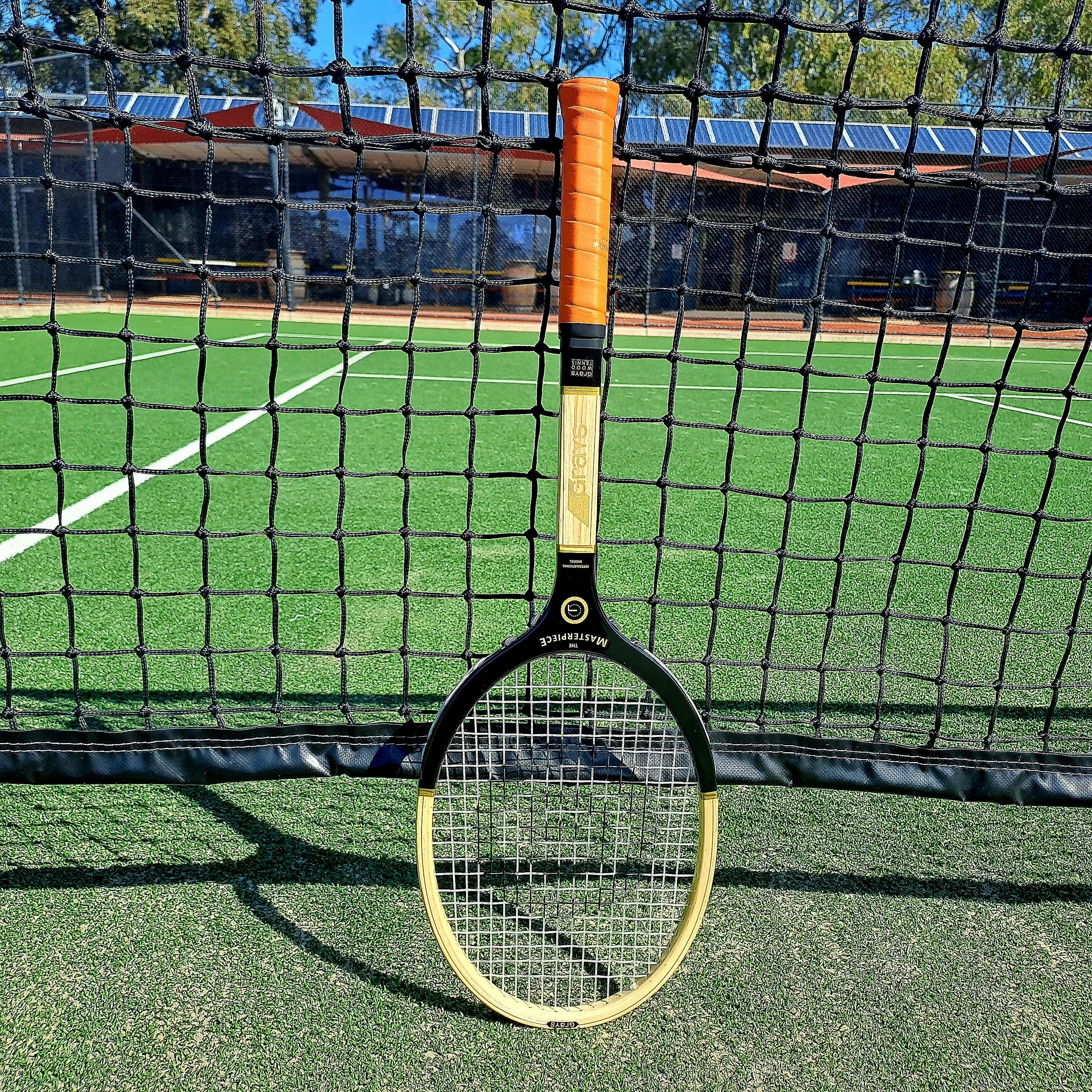 A Grays The Masterpiece wooden racket resting against the net on a grass tennis court with clubhouse in the background.