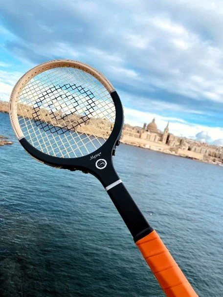 A wooden tennis racket held up against a scenic coastal background with water and buildings.