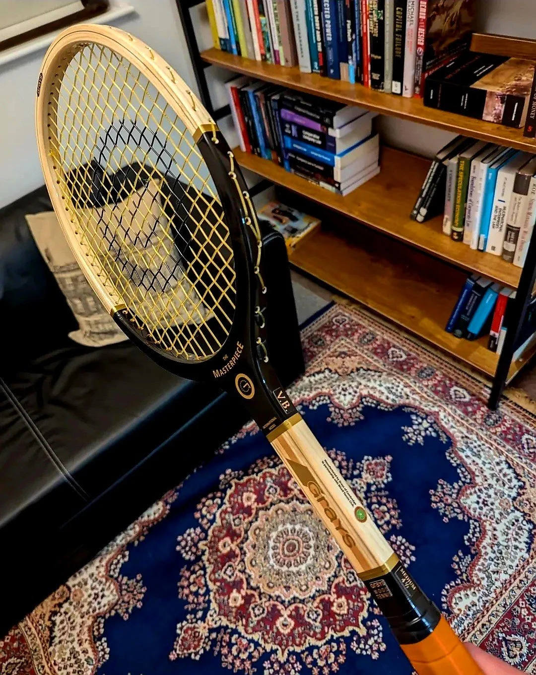 A wooden tennis racket with gold strings is being held over a patterned rug. In the background is a bookshelf filled with books and a black leather couch with a pillow.