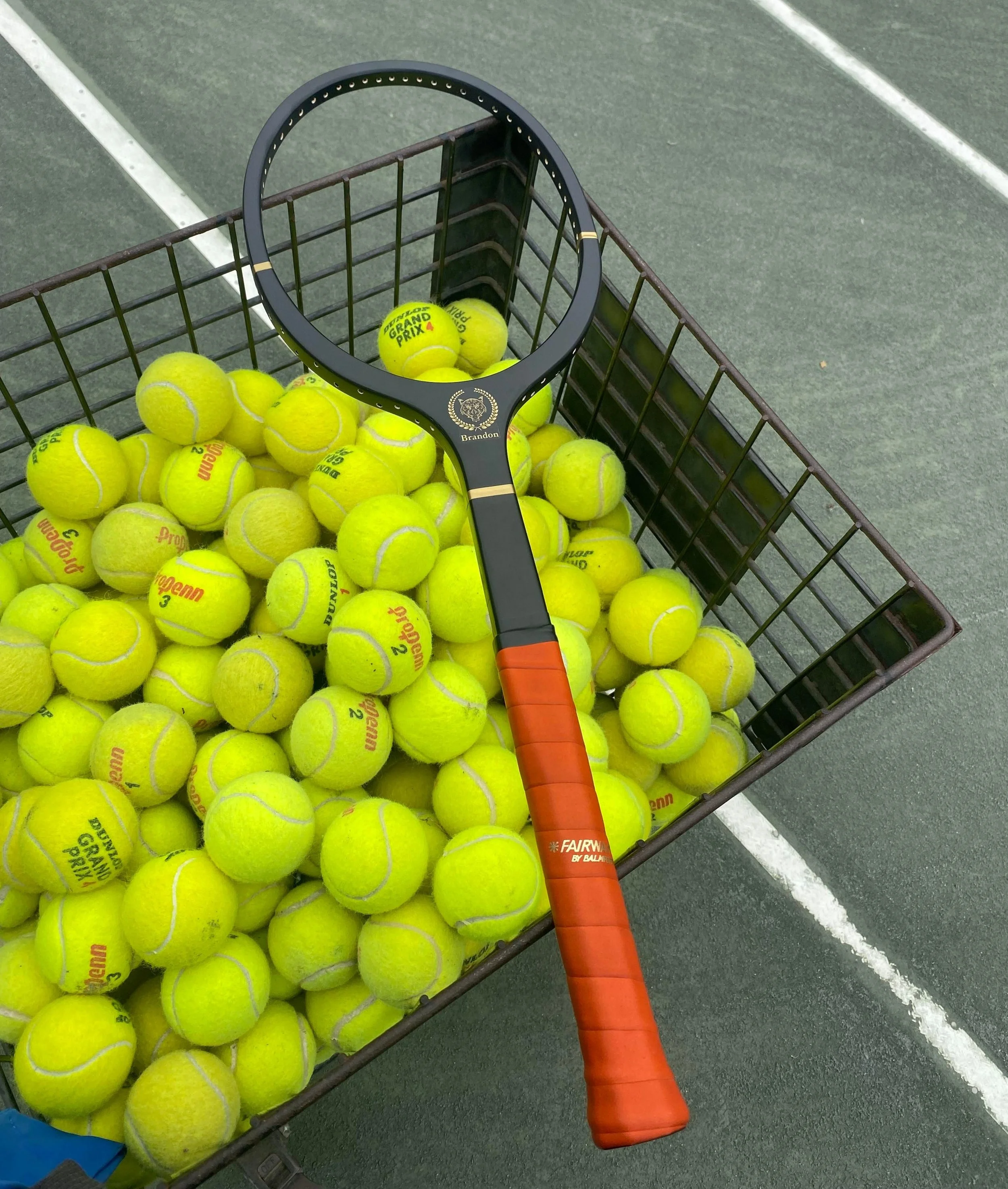 Grays wooden tennis racket with a long brown grip resting on a basket of yellow tennis balls on a tennis court.