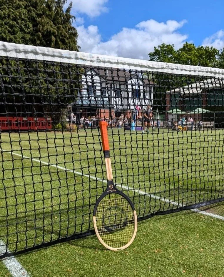 The Masterpiece racket leaning against a net on a grass tennis court.