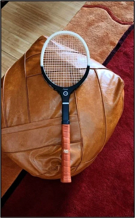 Tennis racket with brown grip on brown leather chair, red carpet background.
