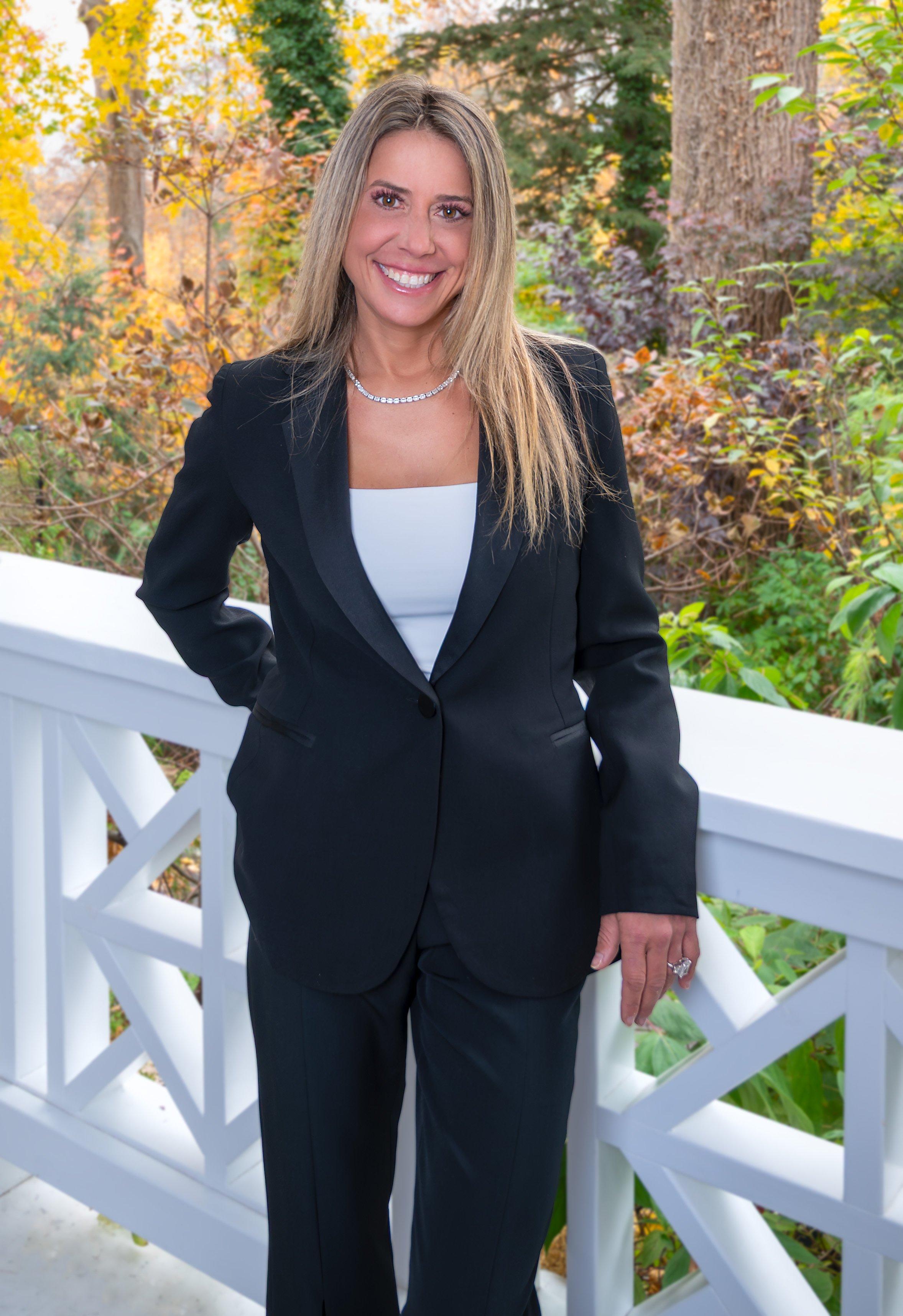 “Professional outdoor headshot of a woman in a black suit smiling while standing on a white railing with autumn foliage in the background.”