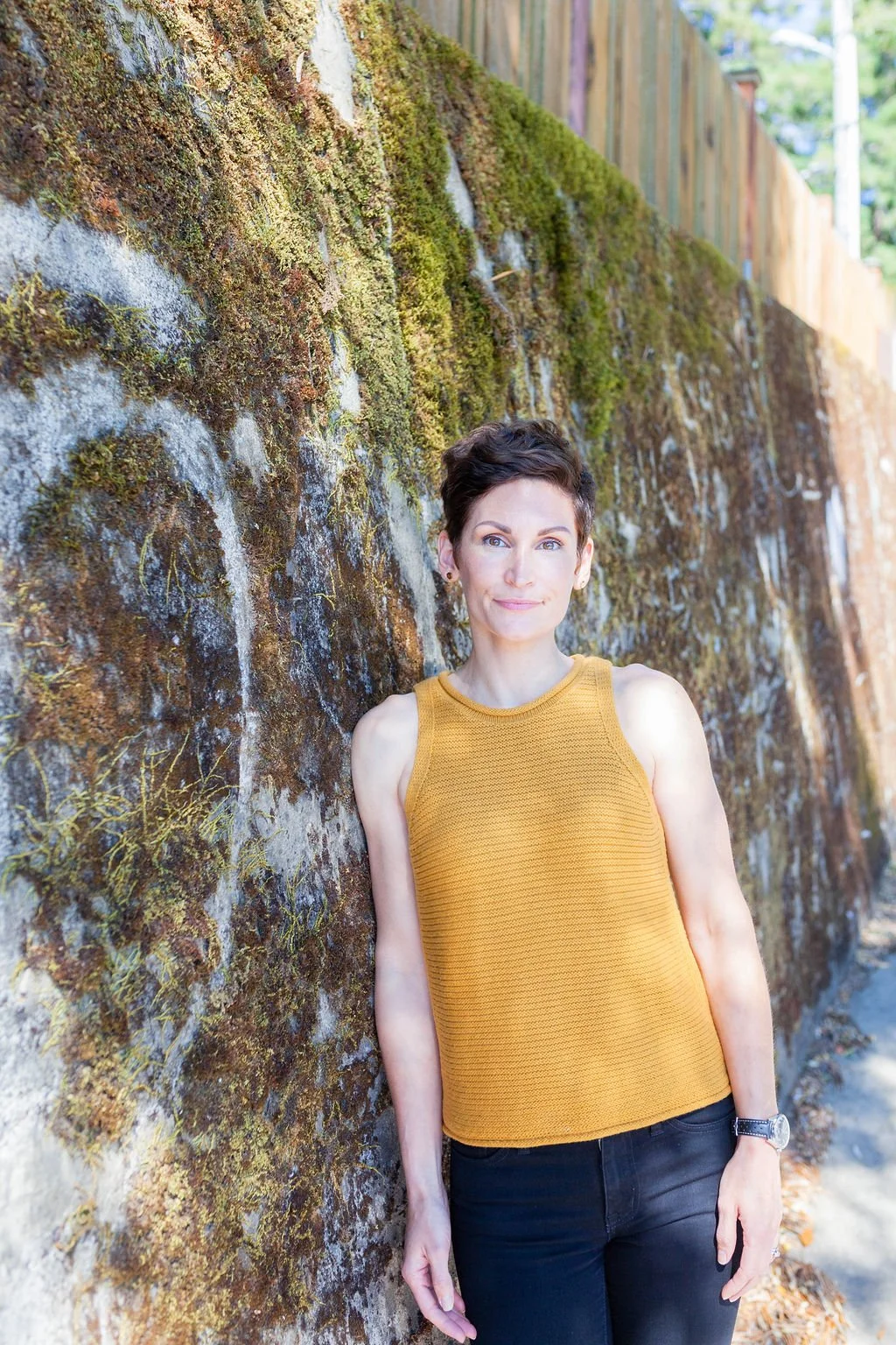 A woman with short dark hair, wearing a sleeveless yellow top and black pants, stands next to a moss-covered brick wall outdoors on a sunny day. Certfied Life Coach Washington.