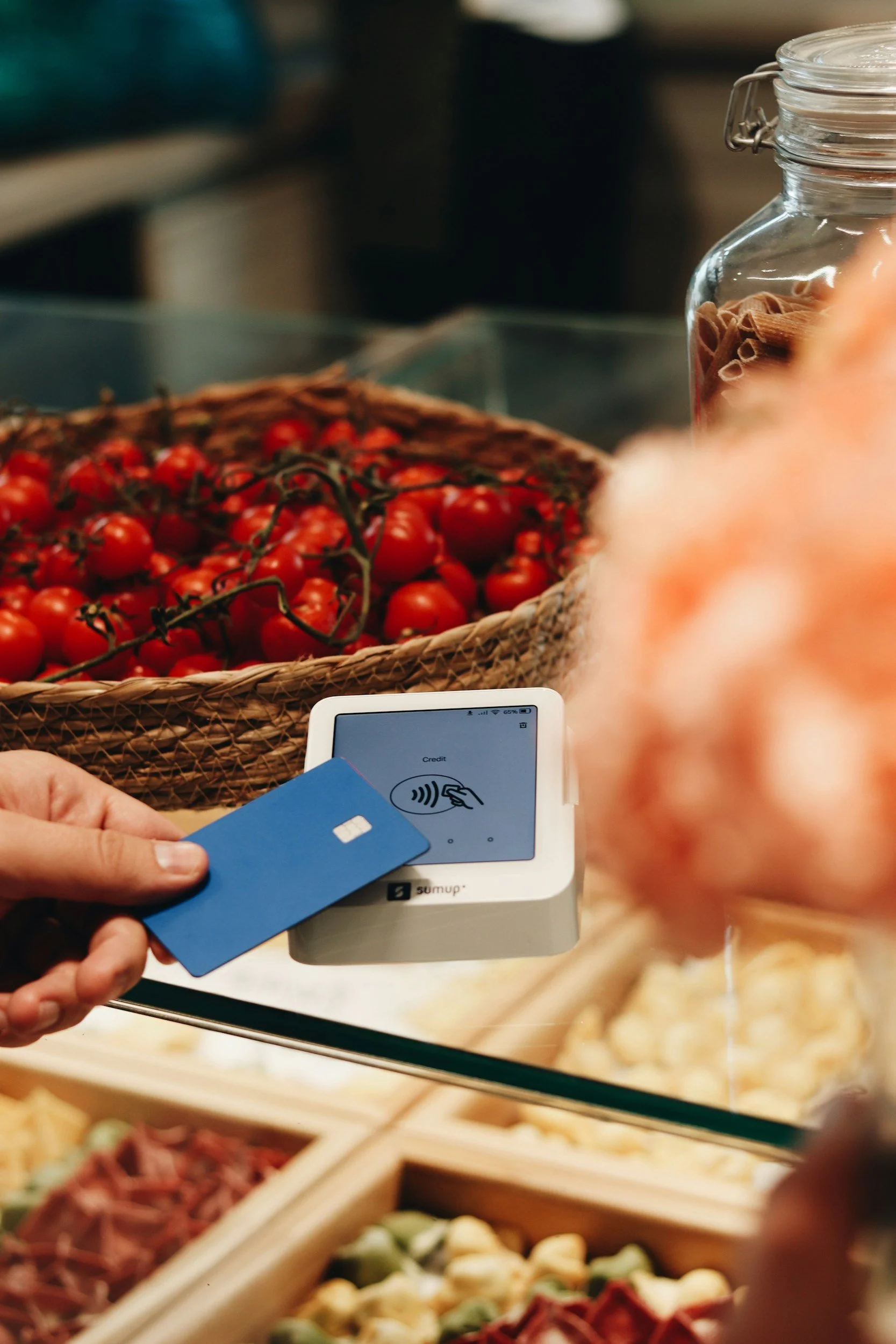 Someone using a contactless payment terminal with a blue credit card at a grocery store, with fresh tomatoes and other produce visible in the background.