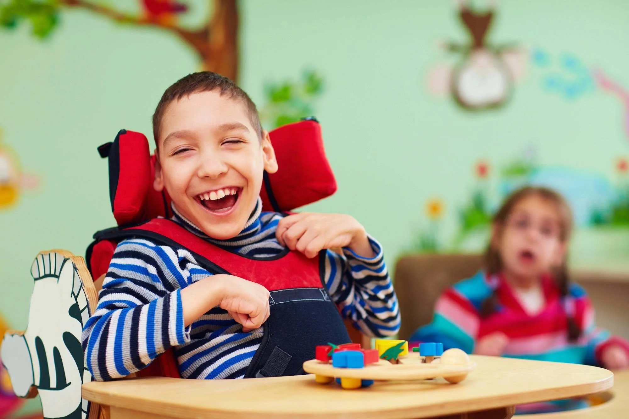 A smiling boy with a disability in a wheelchair, sitting at a table with colorful toys, in a bright room with children playing in the background.
