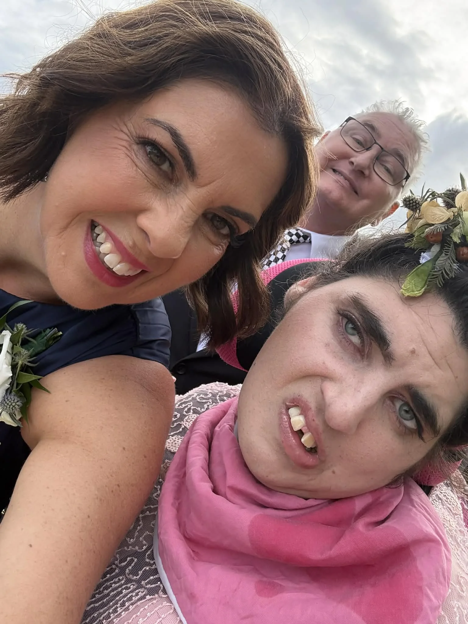 Three people taking a selfie outdoors. The woman in the foreground has short dark hair, a big smile, and is wearing a dark dress with a floral corsage. The other woman has dark curly hair, a pink scarf, and a fierce expression. The man in the background has glasses, gray hair, and a checked tie, smiling slightly.