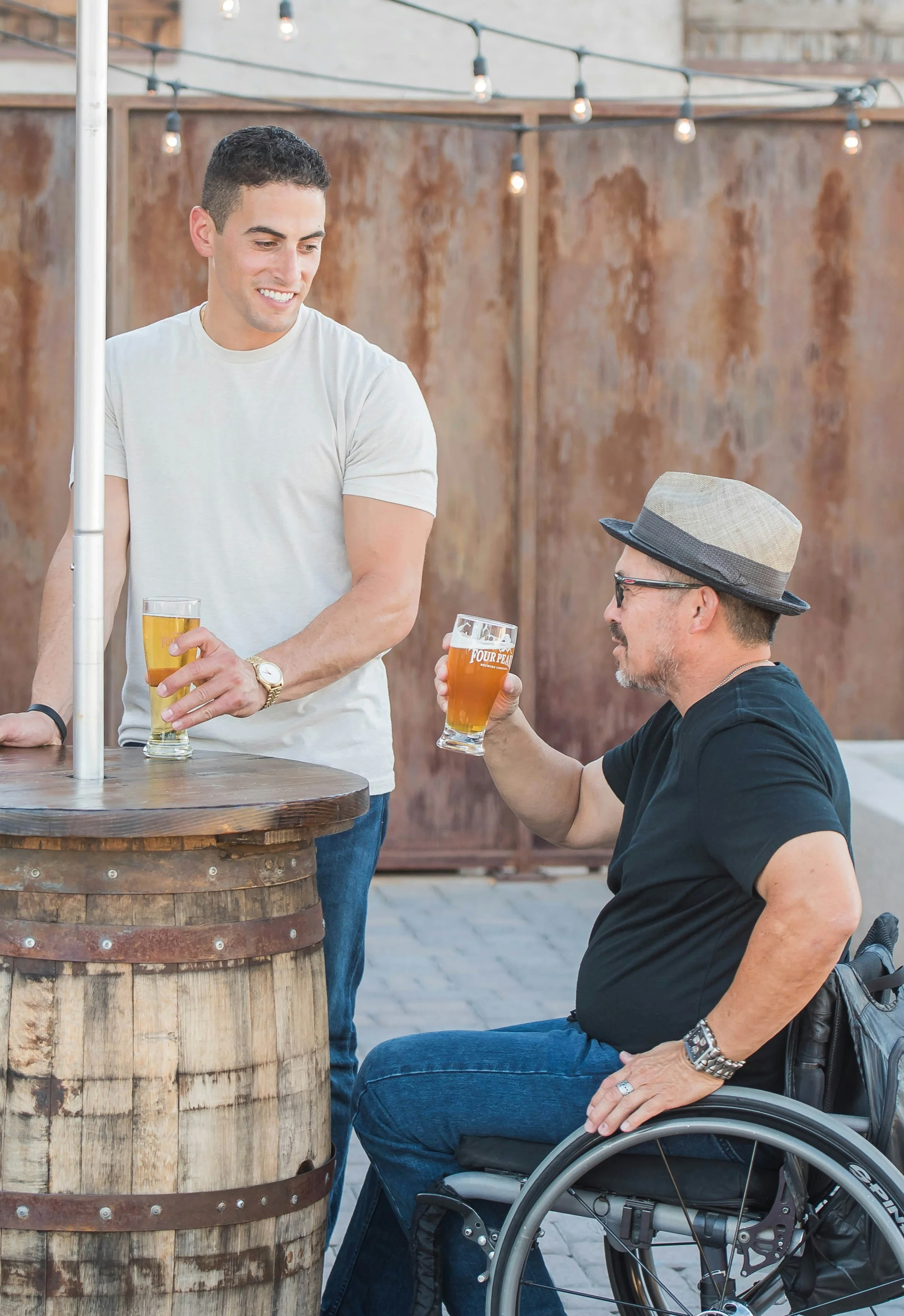 Two men are outdoors, one is standing and the other is seated in a wheelchair. Both are holding glasses of beer. The standing man is smiling and wearing a light-colored t-shirt, while the seated man is wearing a hat, sunglasses, and a dark t-shirt.