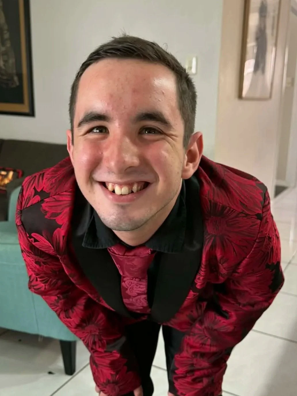 A young man with short dark hair and a big smile showing missing teeth, wearing a red and black floral blazer and matching tie, leaning forward in a room with art on the walls.