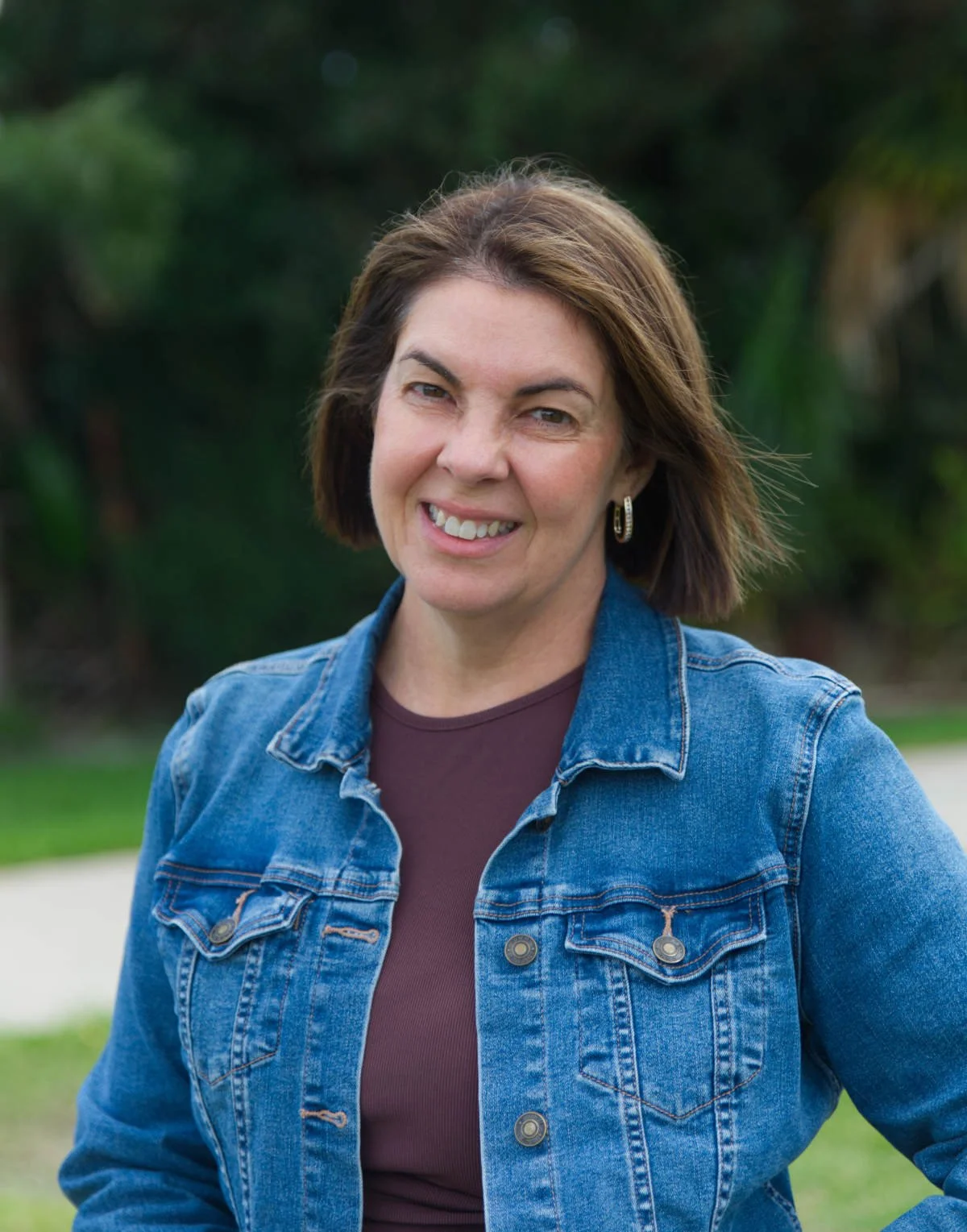 A woman with shoulder-length brown hair, smiling, wearing a denim jacket and a maroon shirt, outdoors with green trees in the background.