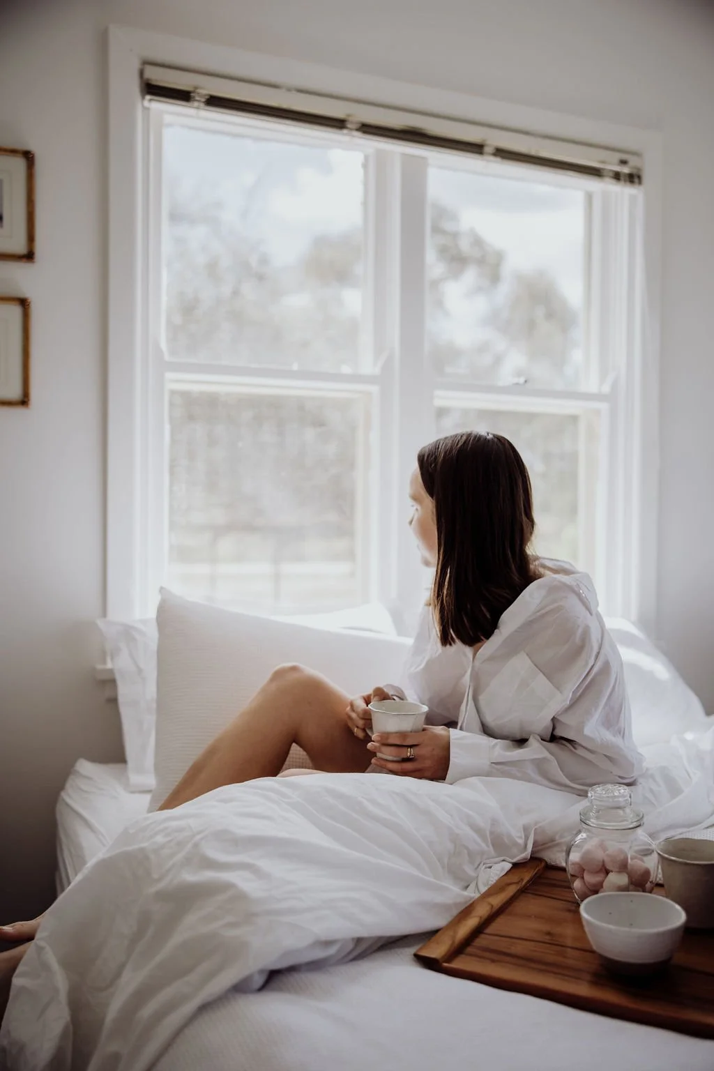 Woman relaxes in bed with a cup of tea looking out north facing windows on the garden