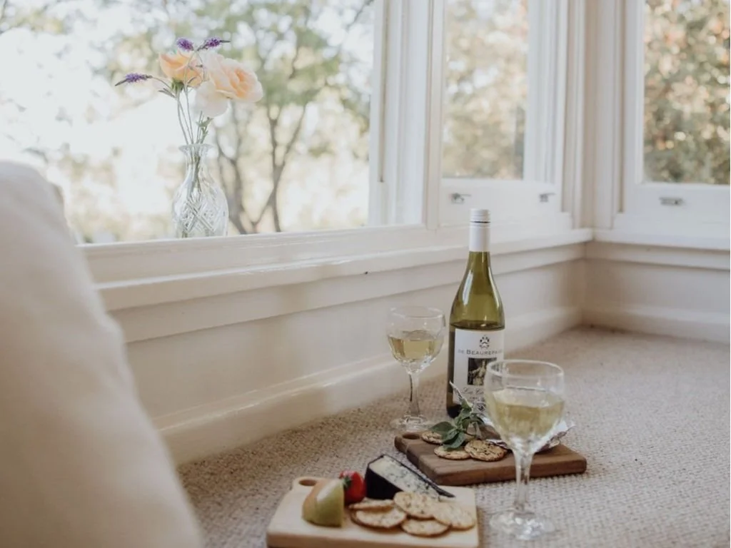 Wine bottle, two wine glasses filled with white wine, and a cheese platter with crackers and grapes on a wooden board, with a vase of flowers on the windowsill.