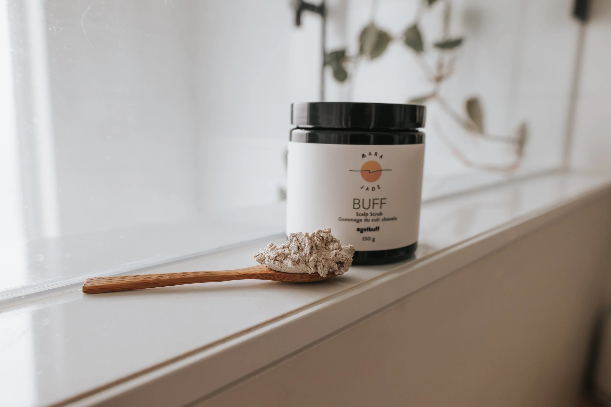A wooden spoon with a heap of gray scalp scrub resting on a white surface, with a black-lidded jar of Marpa Jade Buff scalp scrub behind it, against a blurred background with a plant.