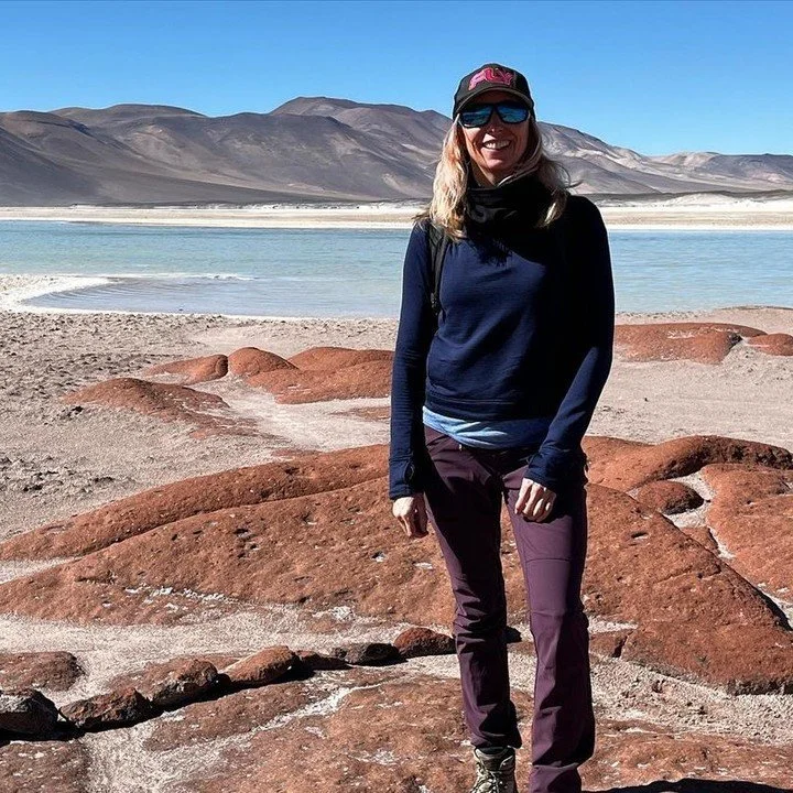 Woman in outdoor gear standing on reddish rocks near a body of water, with mountains in the background under a clear blue sky.
