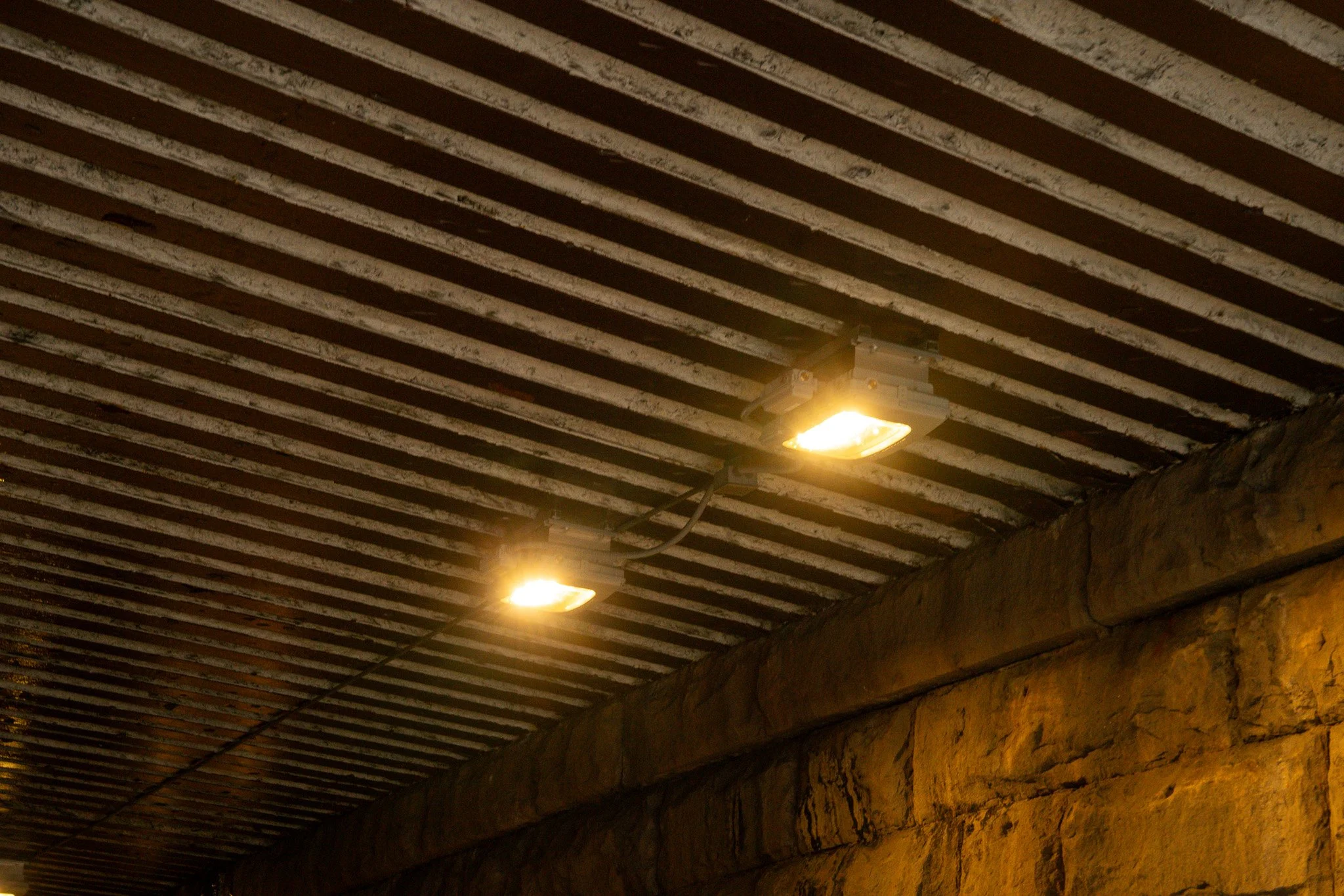 Ceiling of a tunnel with two yellow lights and striped metal beams, stone wall on the side.