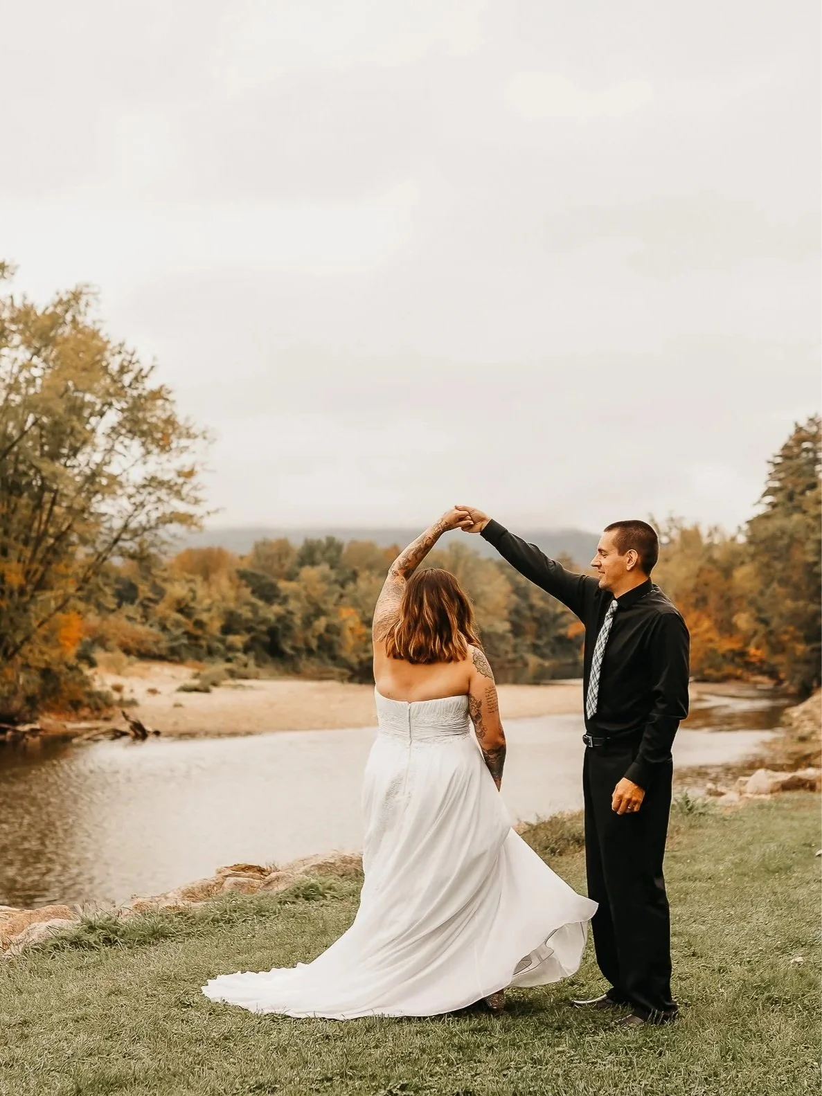 A couple dancing outdoors near a river during fall, with trees in autumn colors in the background. The woman is in a white wedding dress, and the man is in a black shirt and black pants.
