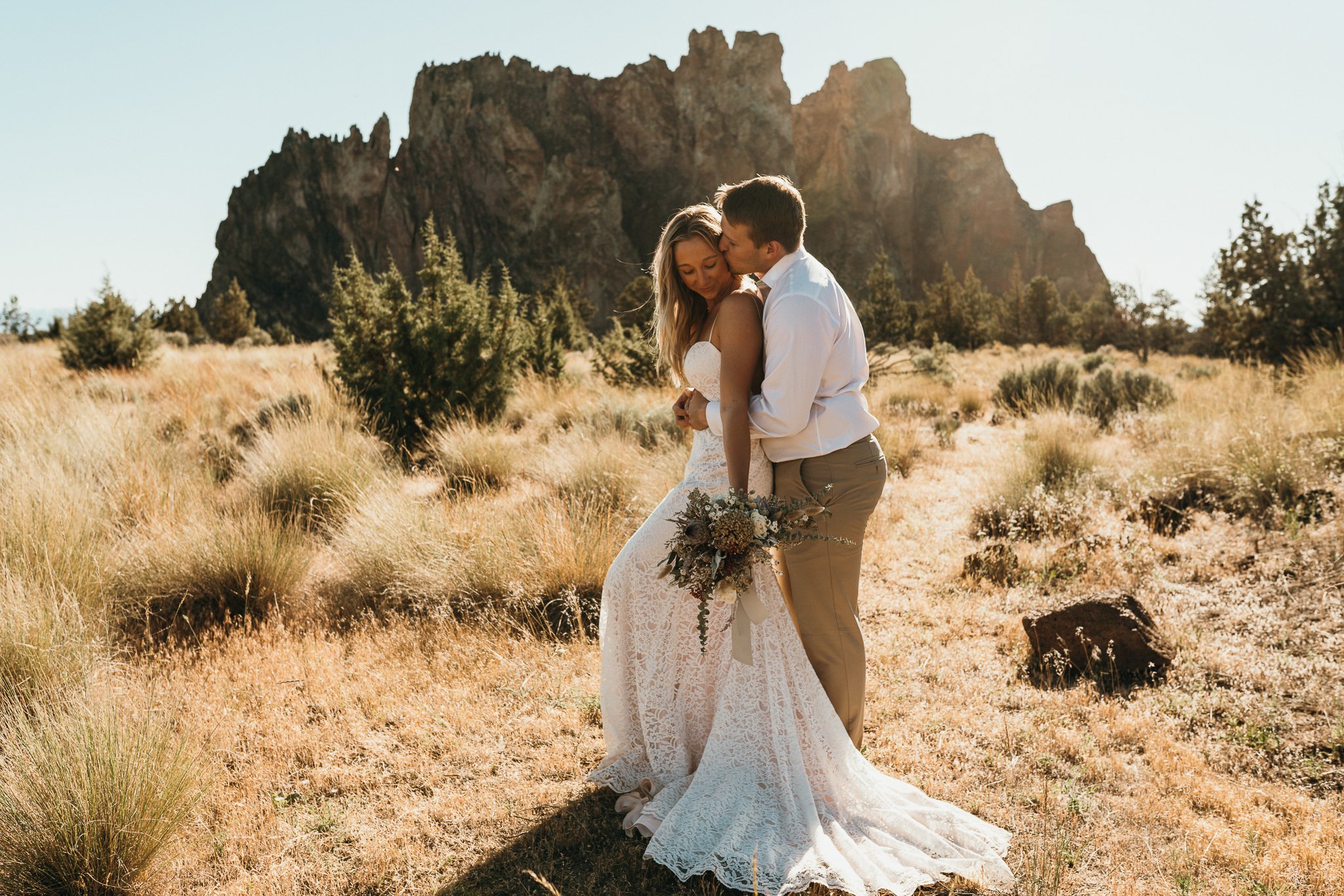 A bride and groom embracing outdoors in a desert landscape with tall rocky formations in the background on a sunny day.