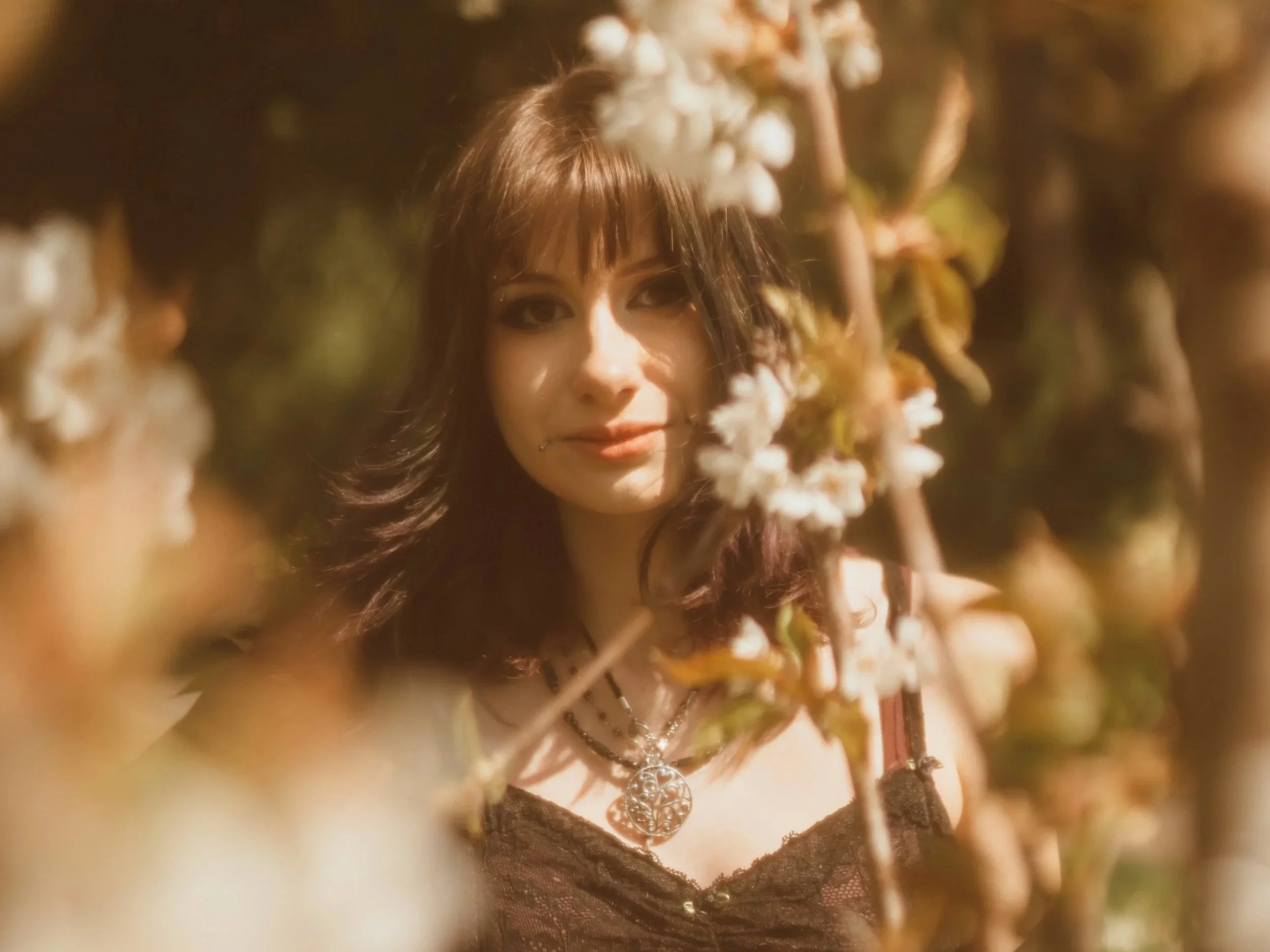 A woman with long brown hair and tattoos on her arm, wearing a black choker and jewelry, standing outdoors among trees and flowers, with sunlight highlighting her face.