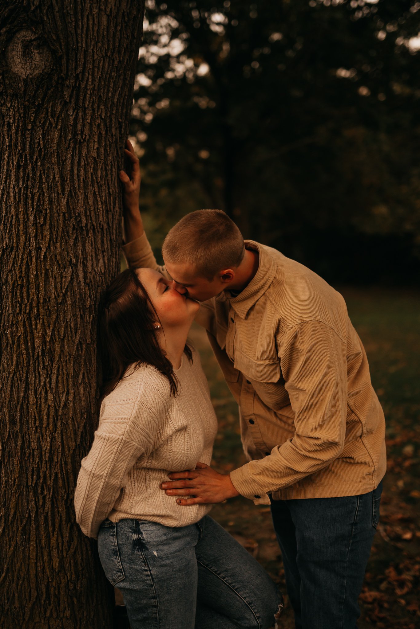 A couple sharing a kiss outdoors near a tree at sunset, with the woman leaning against the tree and the man leaning in to kiss her.