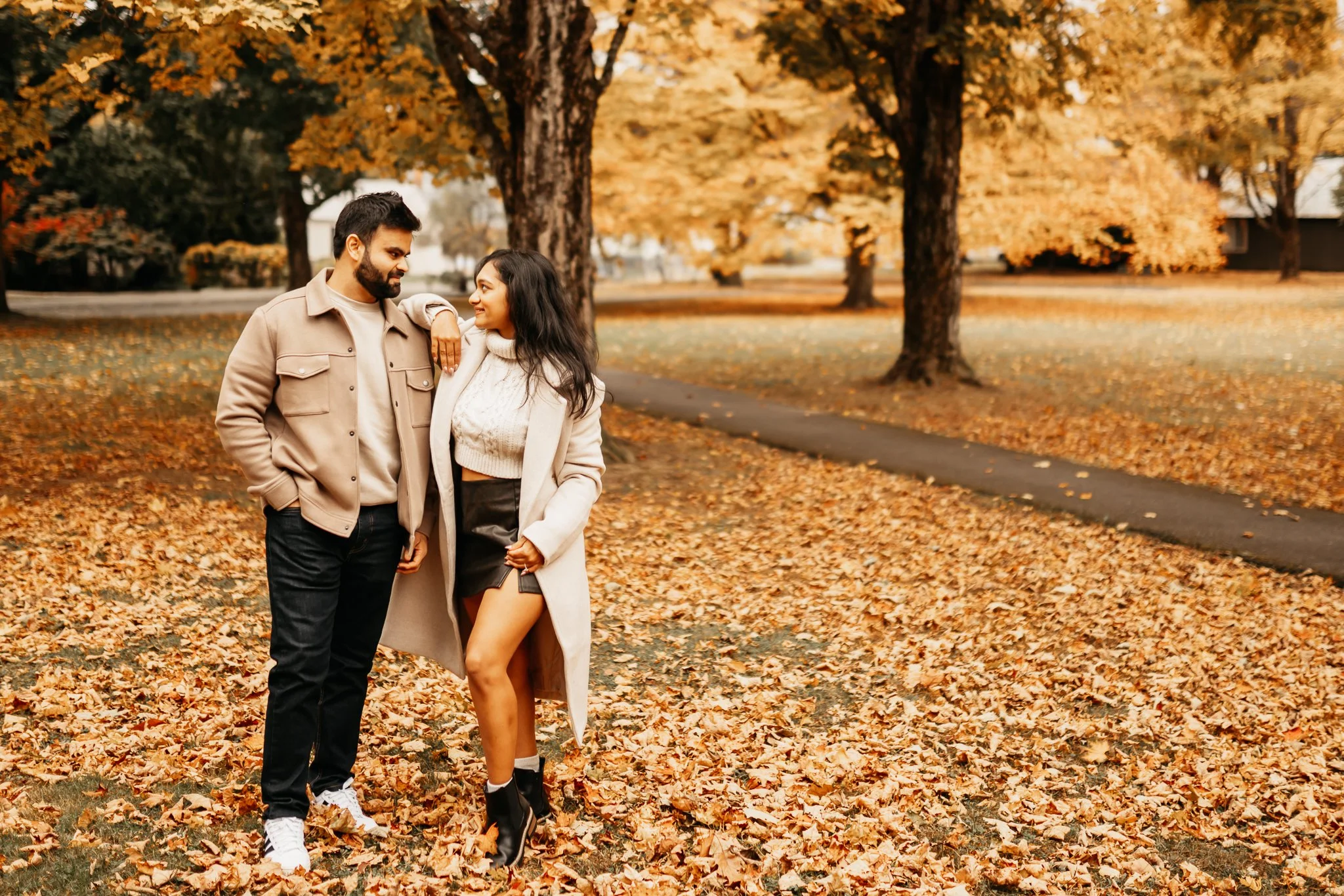 A young man and woman standing together in a park covered with fallen autumn leaves, with trees in the background displaying orange and yellow foliage.