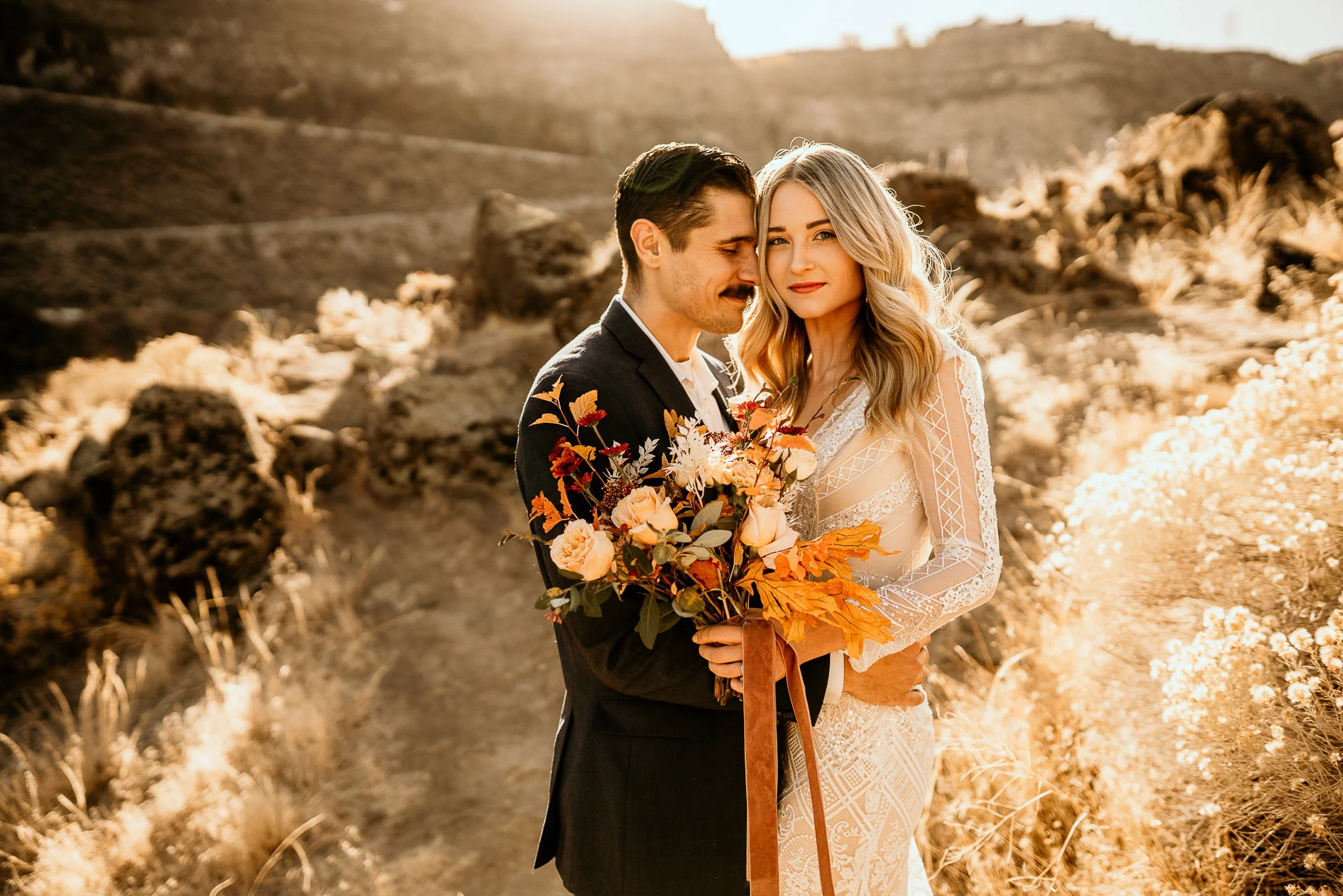 A couple in wedding attire embracing outdoors in a sunlit desert landscape with wildflowers and rocky hills in the background.