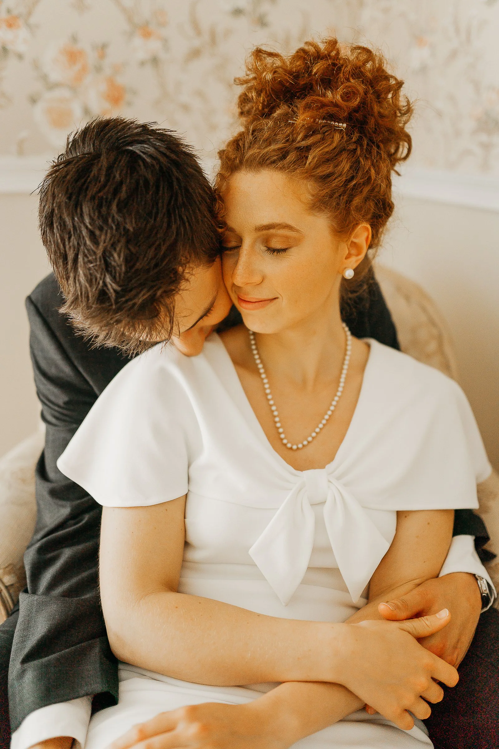 A woman with red curly hair and pearl jewelry smiling with eyes closed, held by a man with short dark hair, on a beige couch in a warmly lit room.