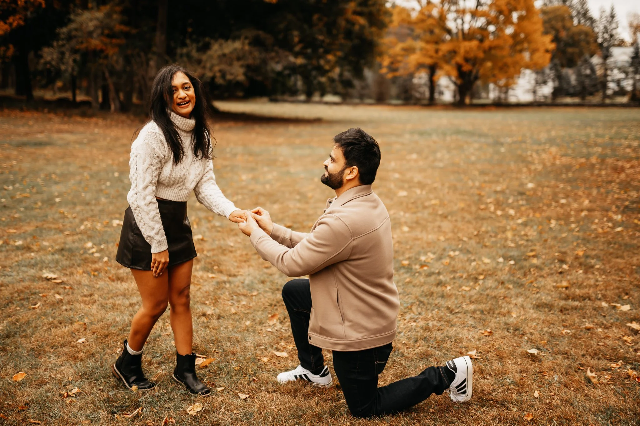 A man proposes marriage to a woman in a park with autumn trees, with the woman smiling happily and holding his hands.