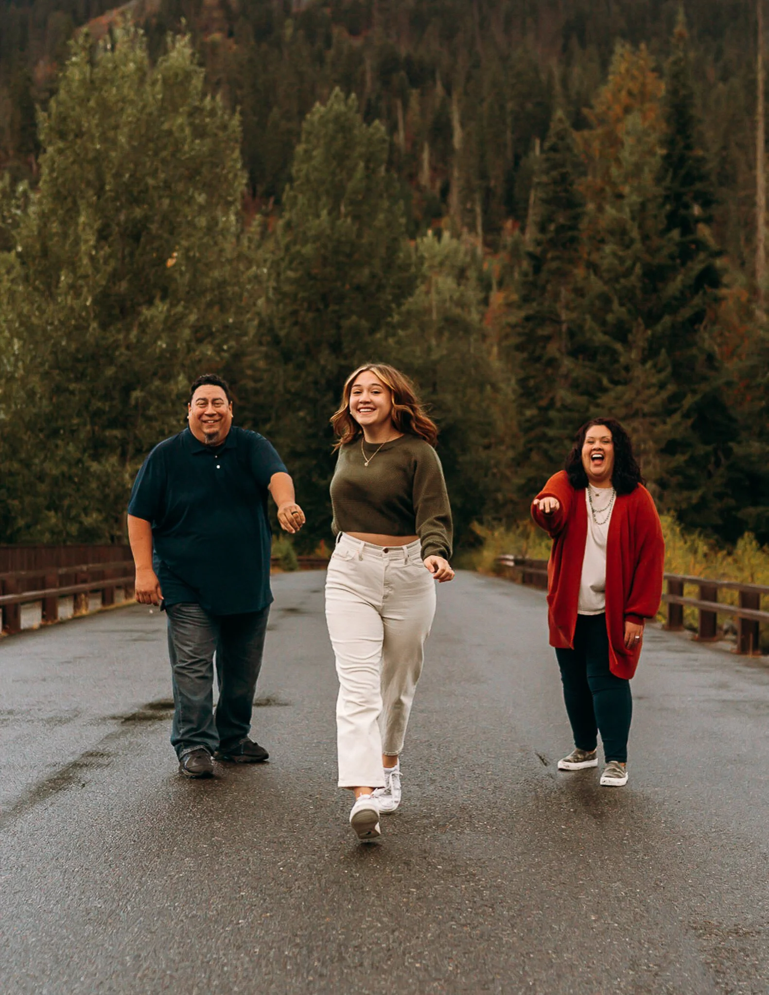 Three people, two women and one man, walking on a wet road in a forested area, smiling and pointing at the camera.