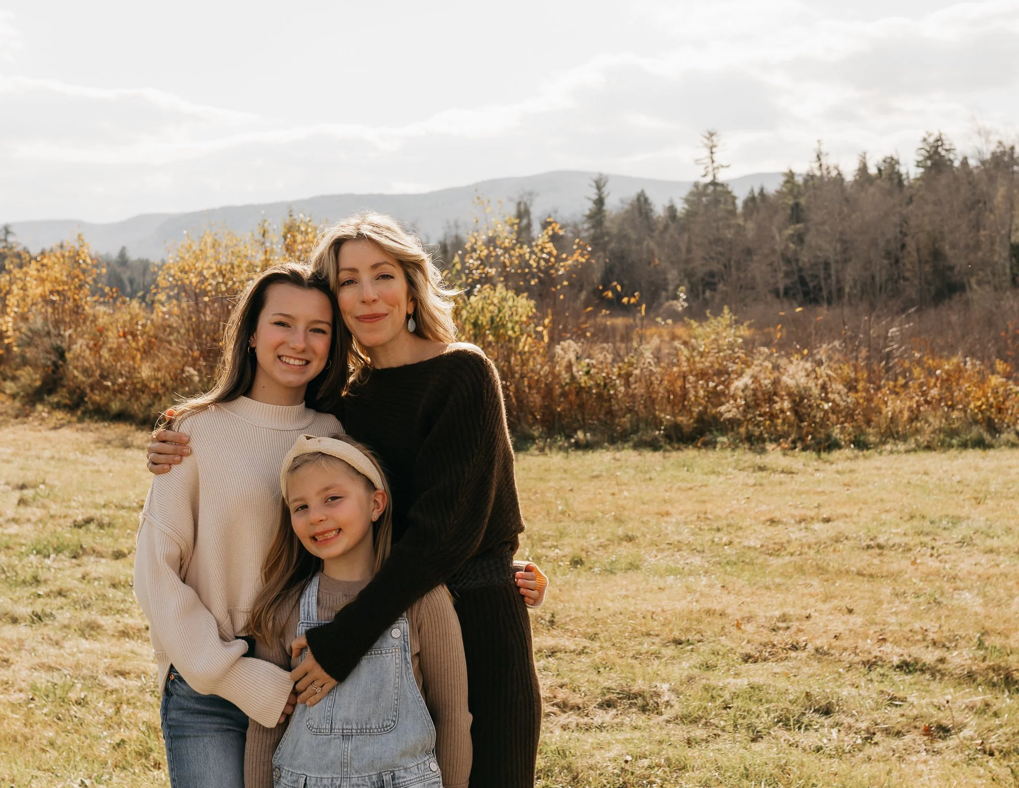 Three females, likely a family, smiling outdoors in a field during autumn, with trees and mountains in the background.