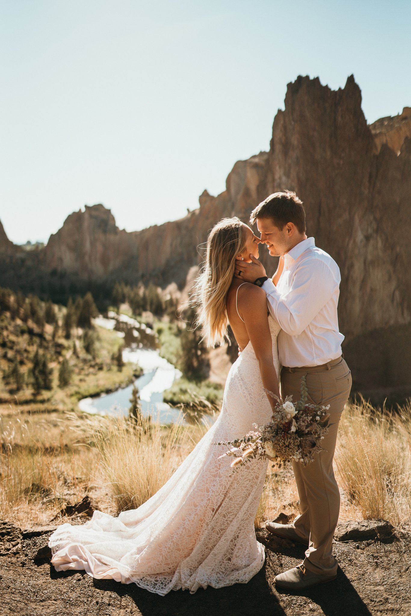 A couple in wedding attire shares a kiss in a scenic outdoor landscape with mountains and a river.
