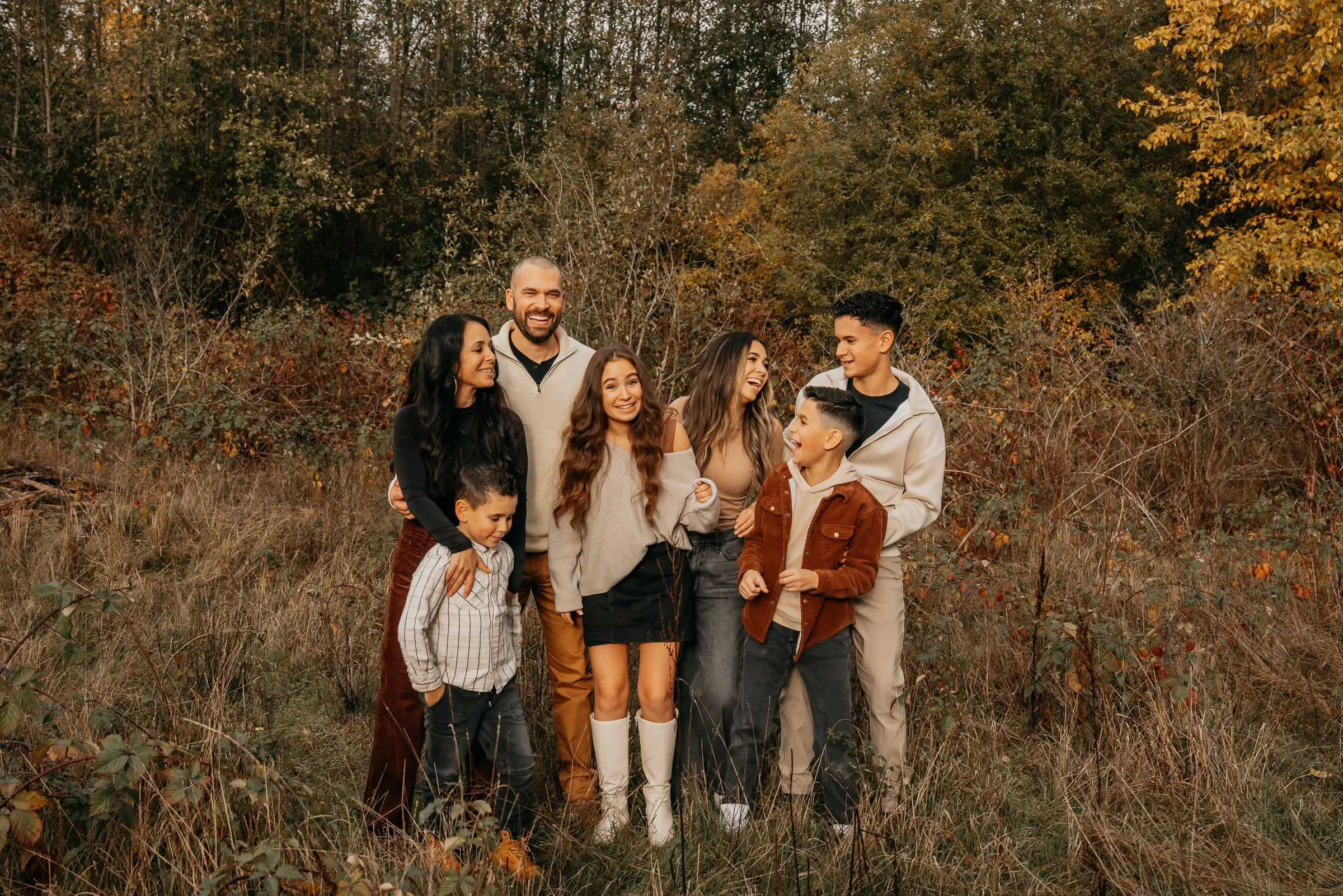 A diverse group of nine people, including adults and children, standing outdoors in a forest during autumn, smiling and enjoying each other's company.