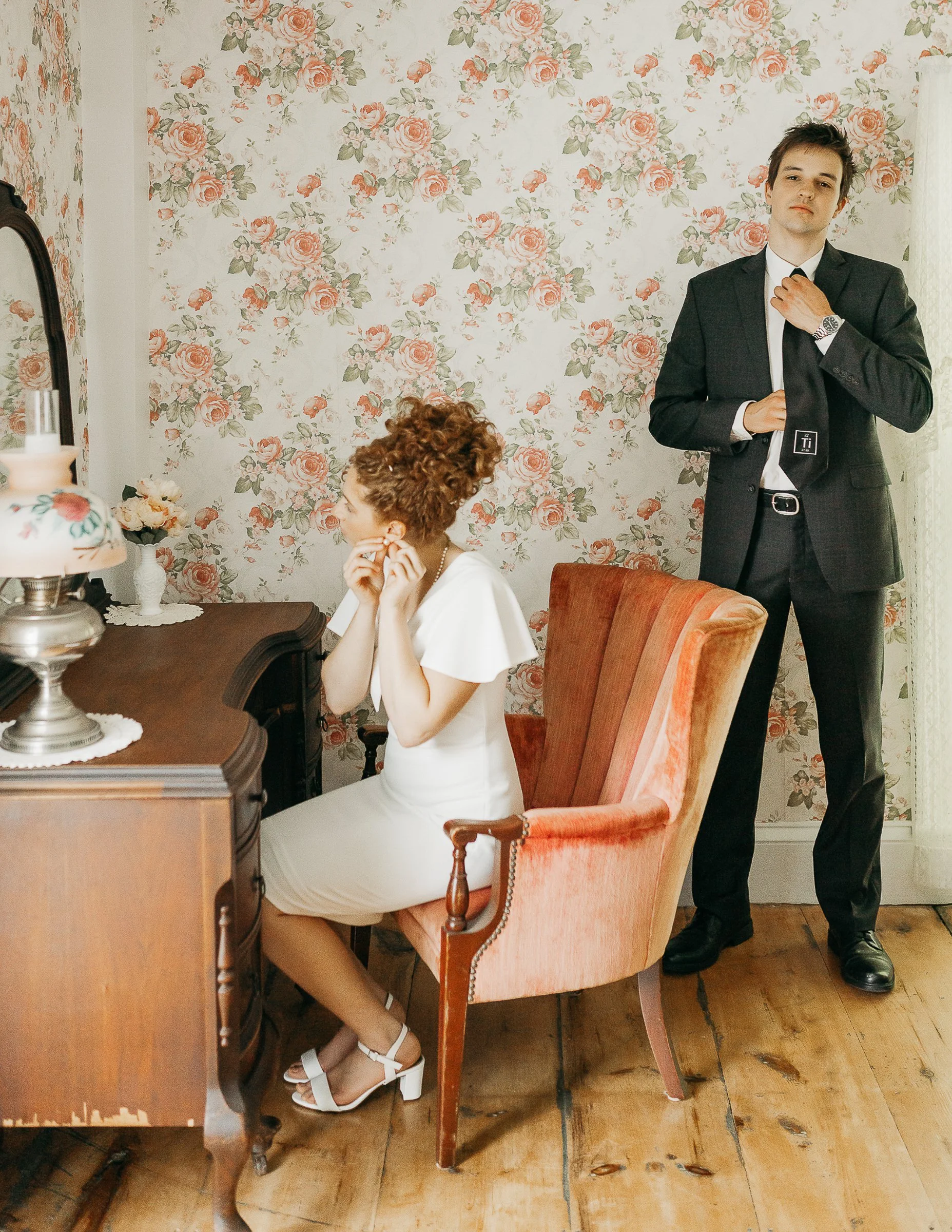A woman in a white dress sitting at a vanity table with a woman in a white dress sitting in a chair, and a man in a black suit adjusting his tie in a room with floral wallpaper.
