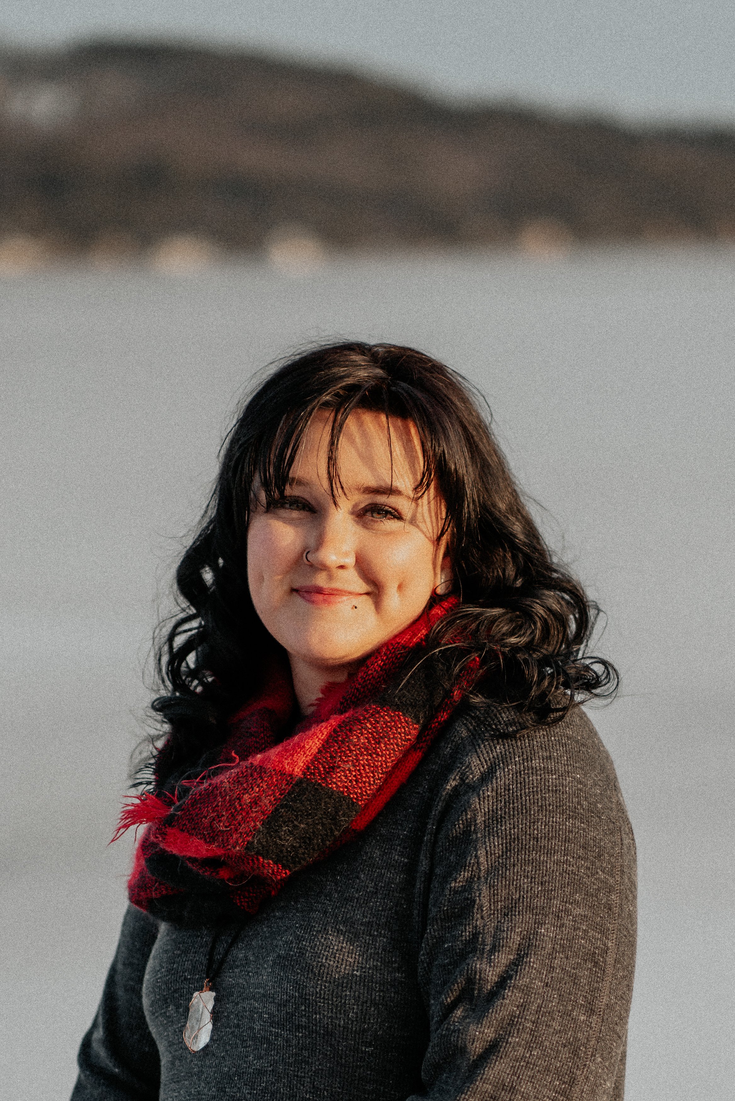 A woman with black curly hair, wearing a grey sweater and a red and black plaid scarf, smiling outdoors during sunset.