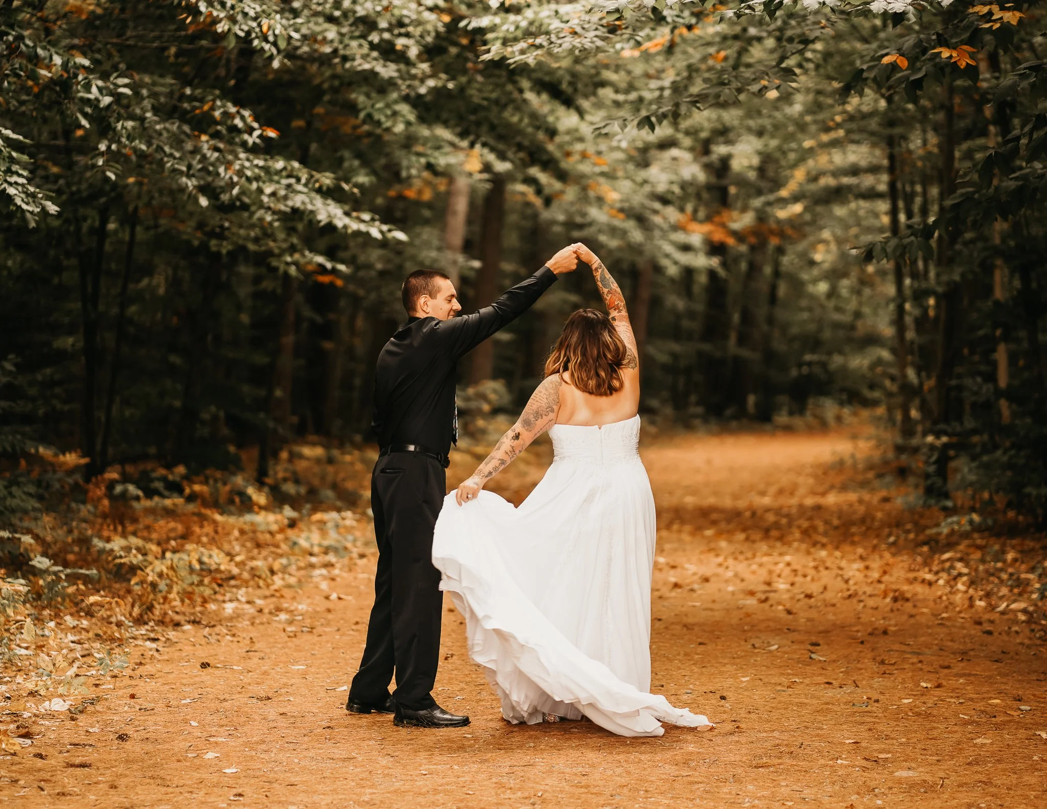 A couple dancing in a wooded area, with the man in a black outfit and the woman in a white dress, holding hands and twirling on a dirt path surrounded by trees.