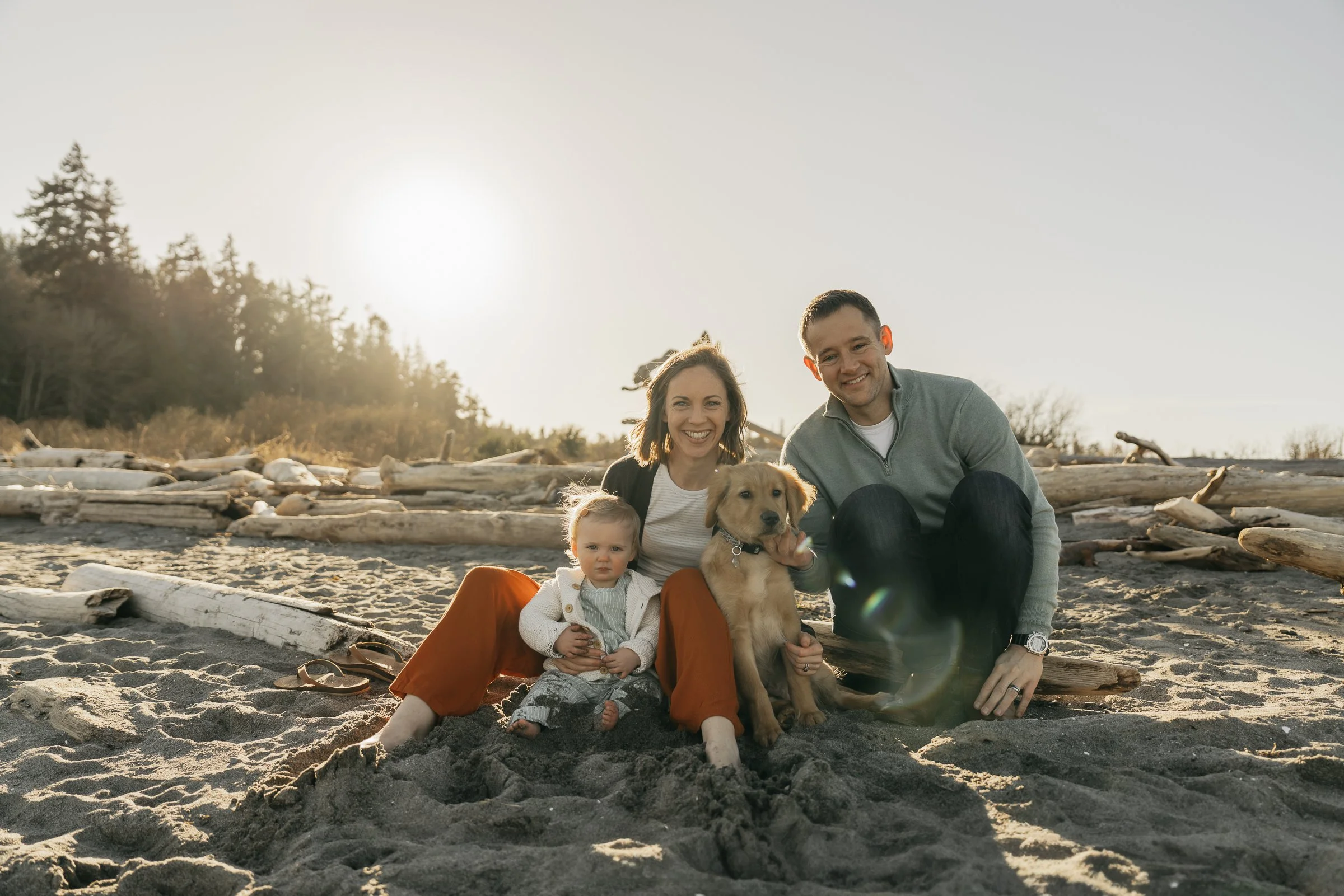 A family of four, including two young children, a woman, a man, and a dog, sitting on a beach with driftwood and trees in the background during sunset.