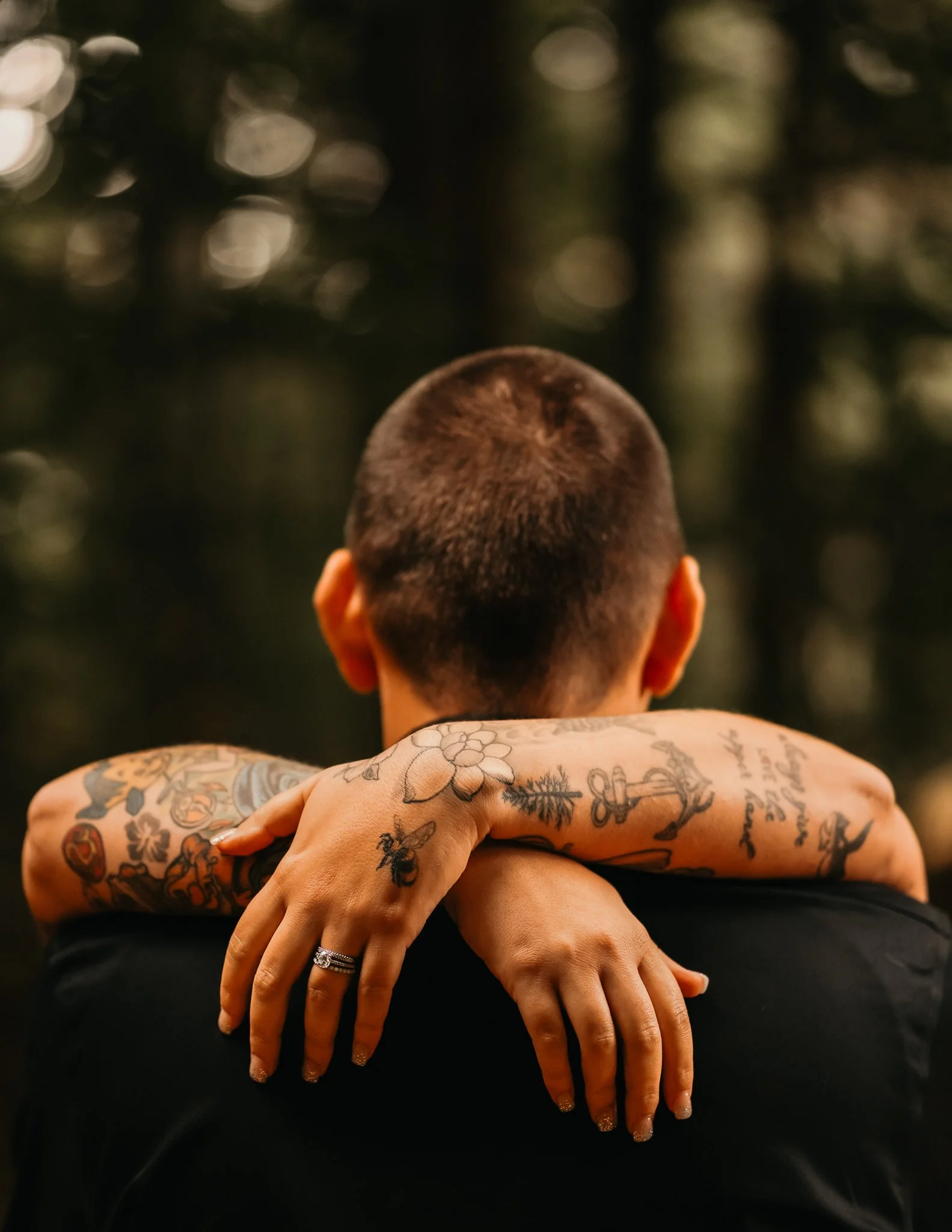 A person with short hair seen from behind, being hugged by a woman with tattoos on her arms and hands, in an outdoor setting with blurred trees in the background.