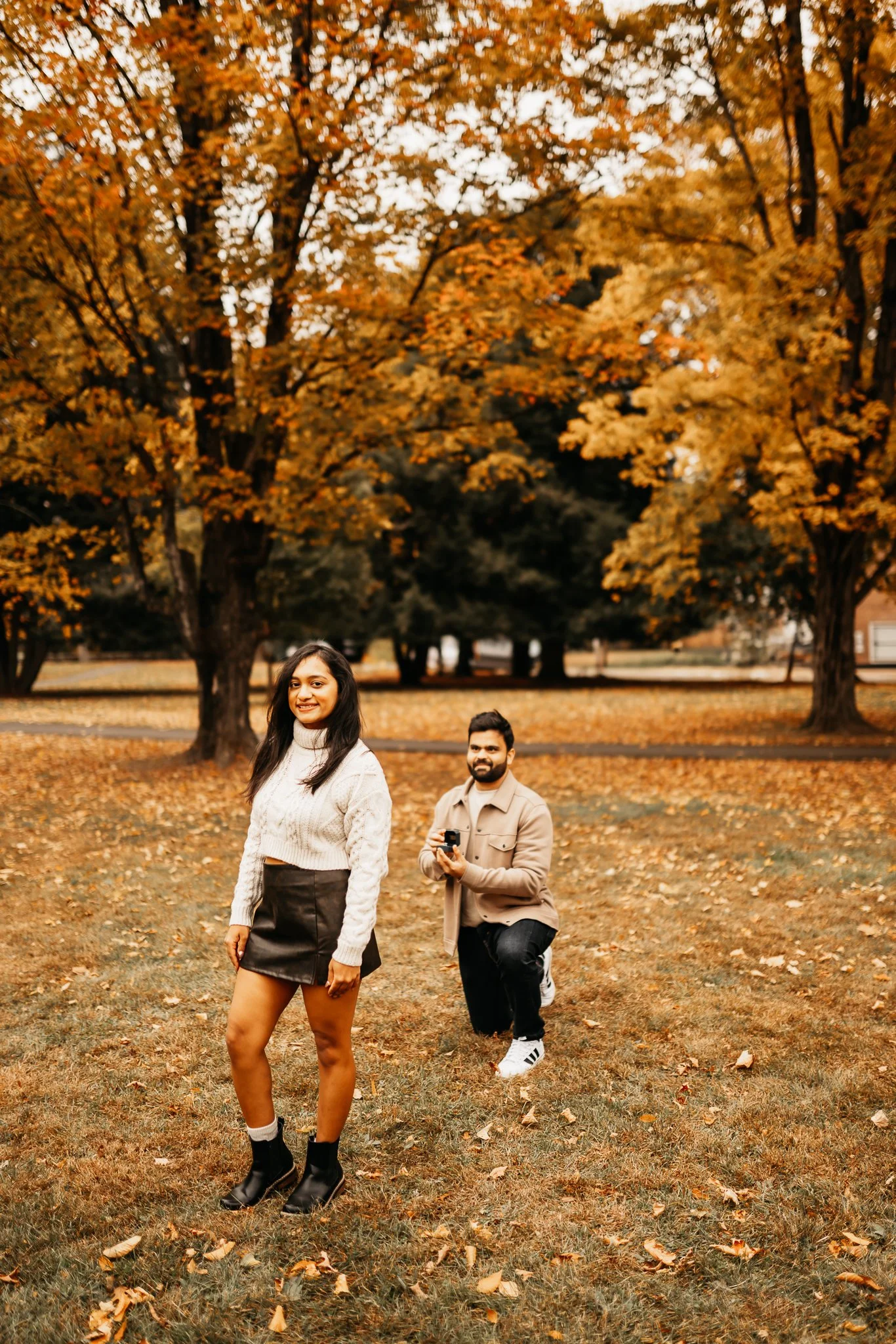 A woman in a white sweater and black skirt standing in a park with autumn orange and yellow trees, while a man kneels behind her holding a camera, both smiling.